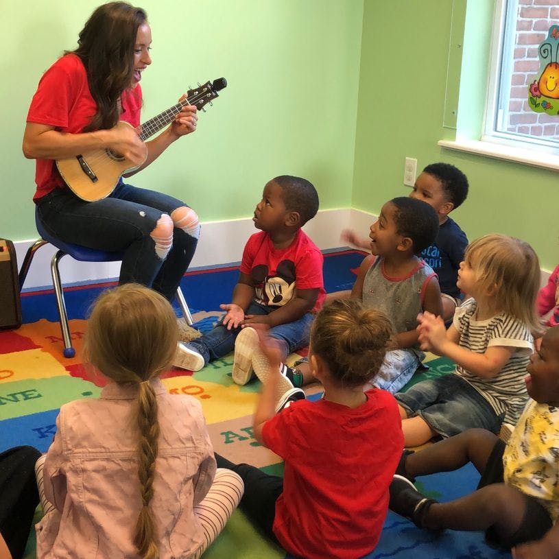 Person playing a ukulele for a group of children sitting on the floor