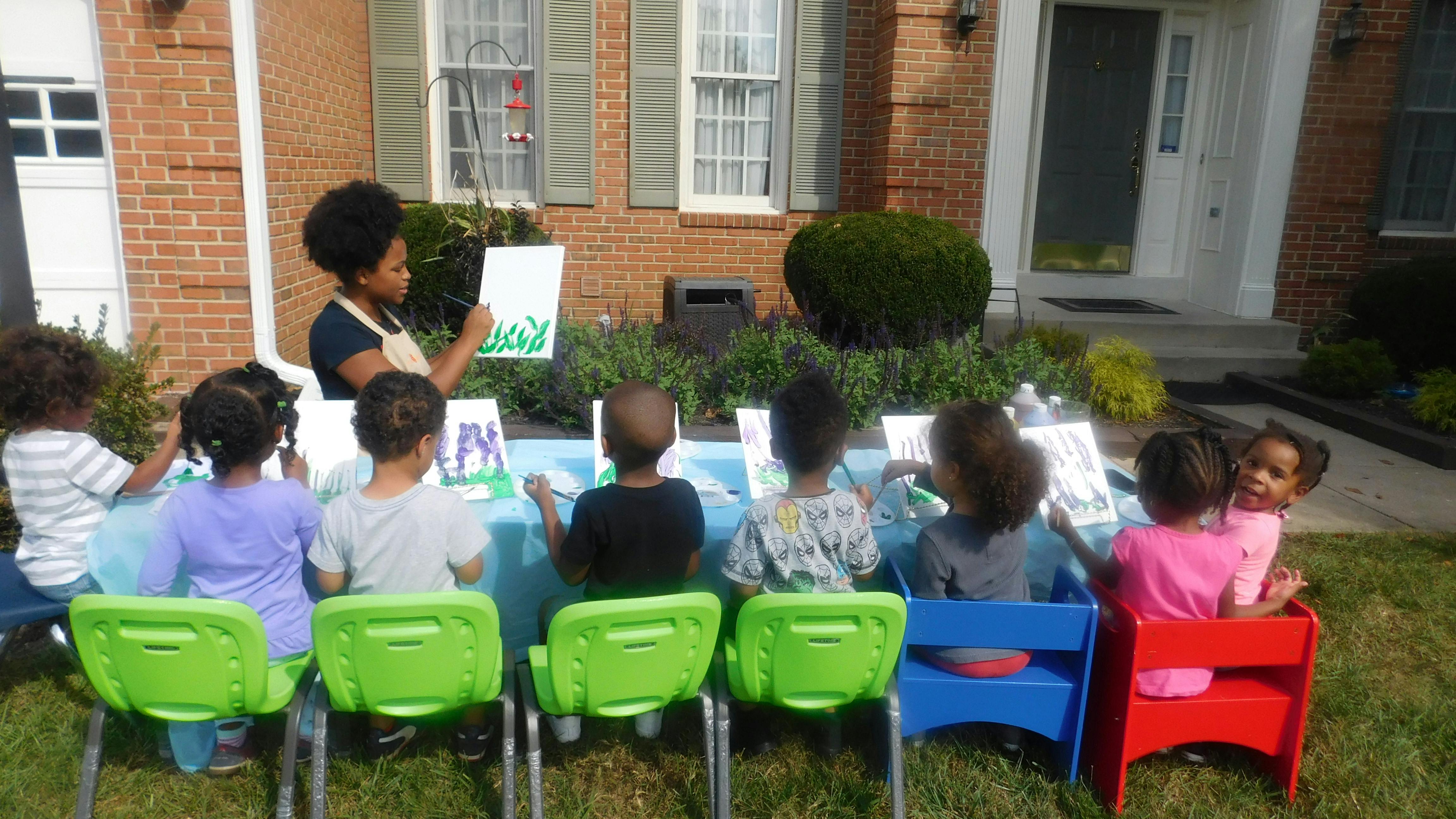 Teacher demonstrating painting to a group of children at a table outdoors
