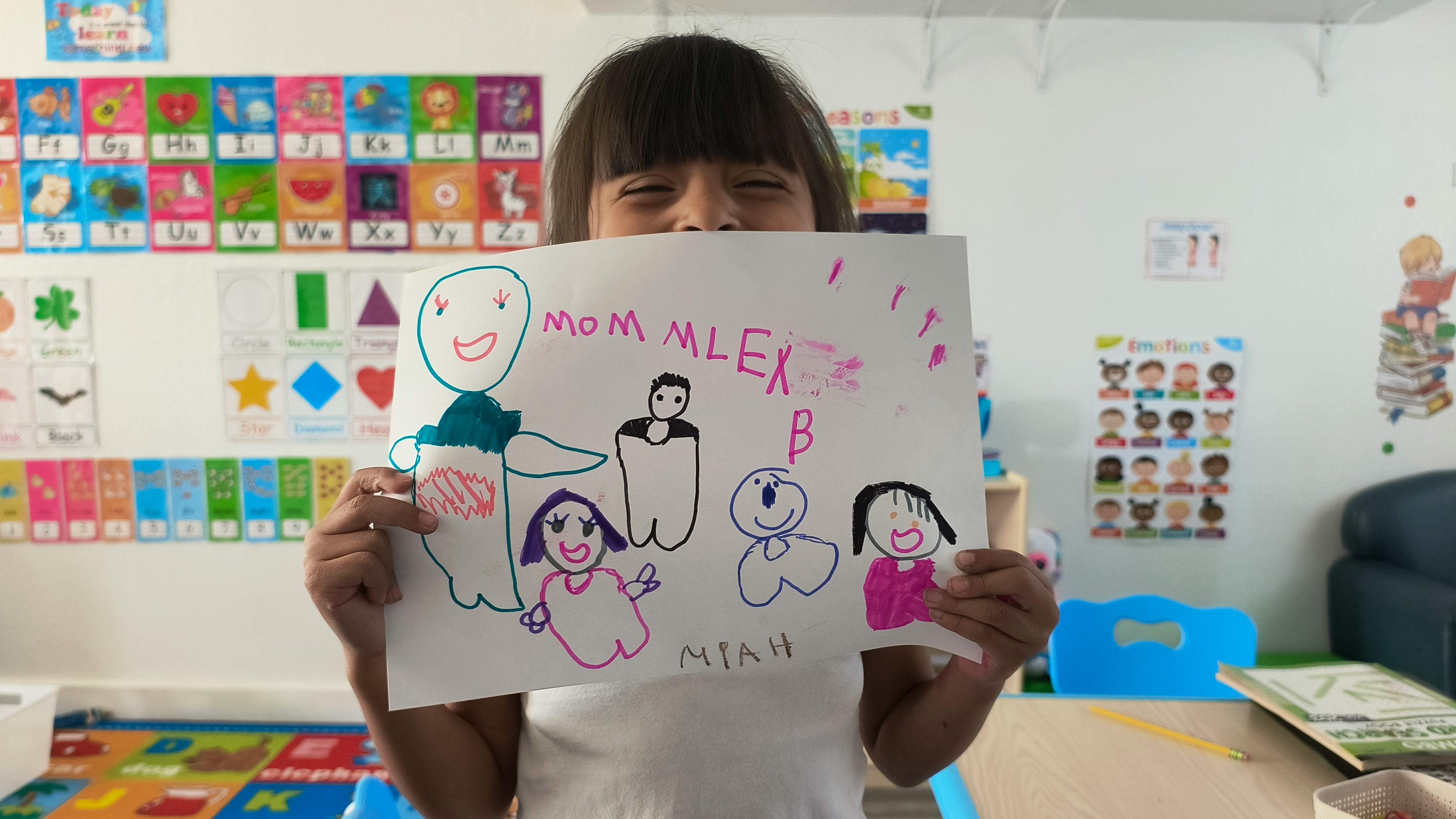 Child holding up a drawing of stick figures with text "Mom Alex B"
