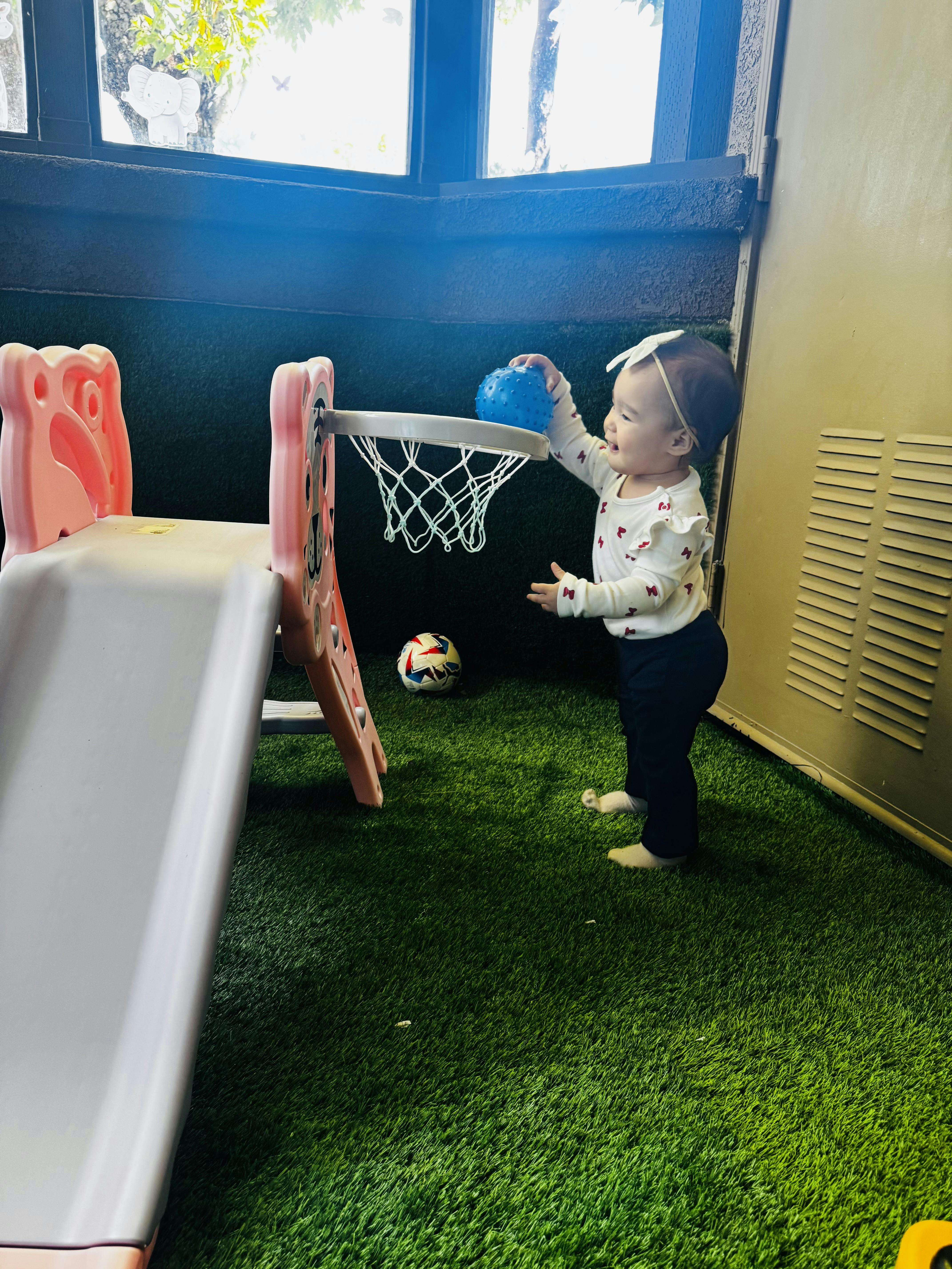 Child playing with a blue textured ball at a small pink basketball hoop