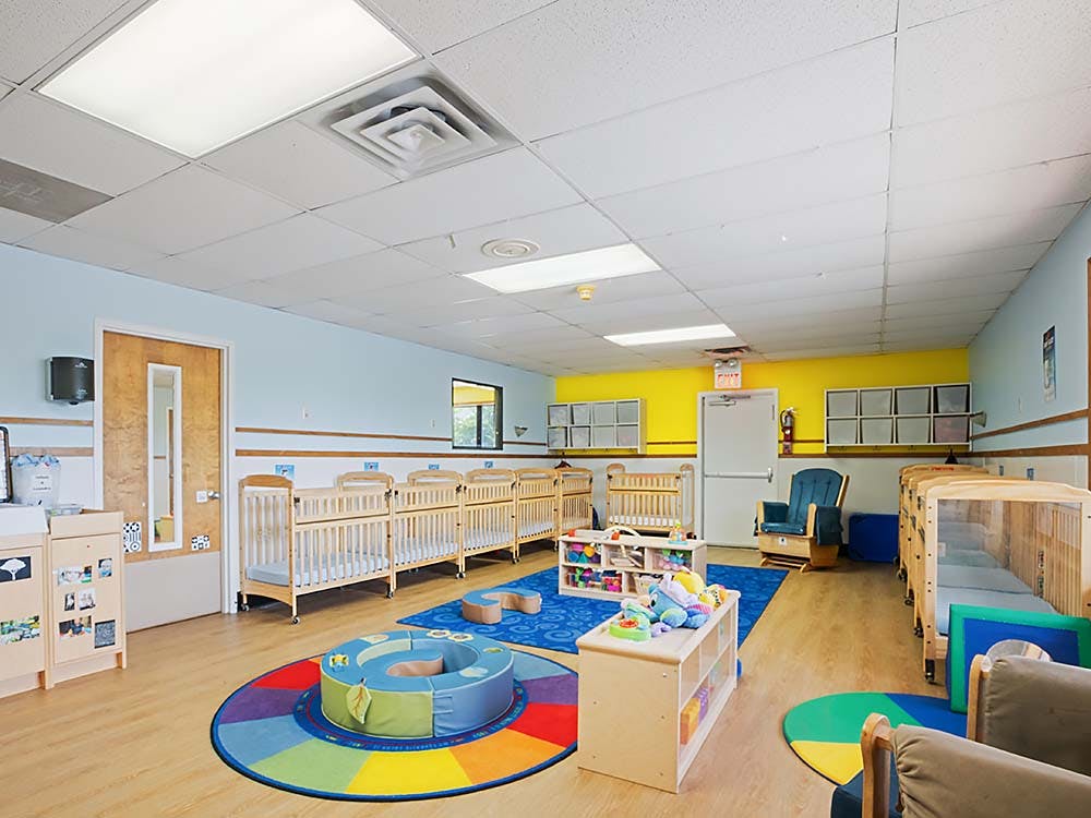 A brightly lit nursery room with multiple cribs, a play mat, and toys