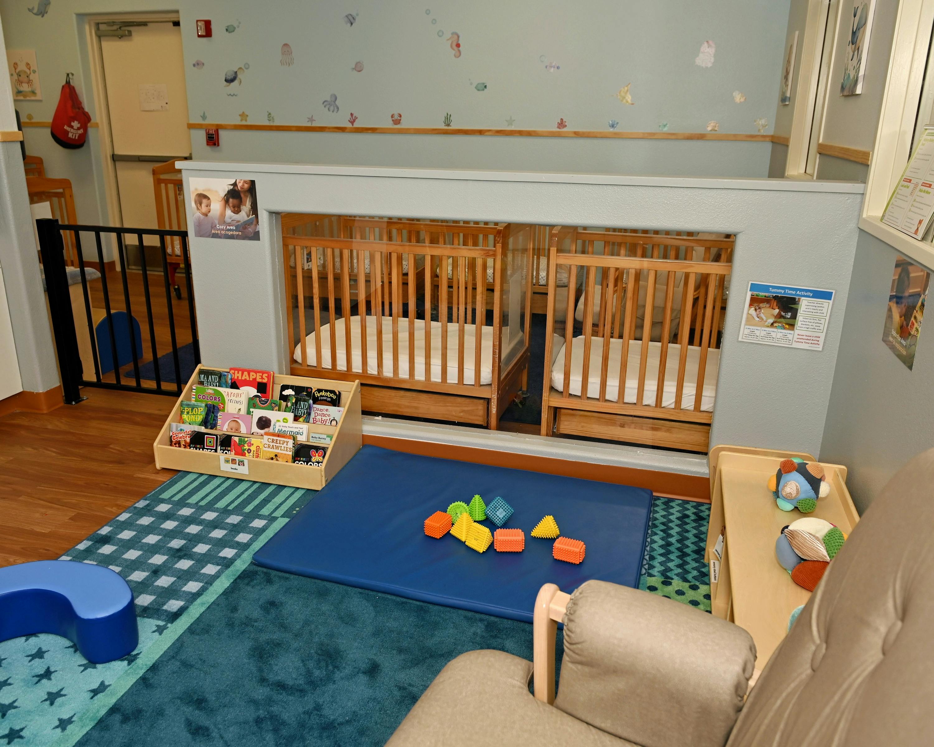 Cribs behind a half-wall in a childcare room with playmats and books on the floor