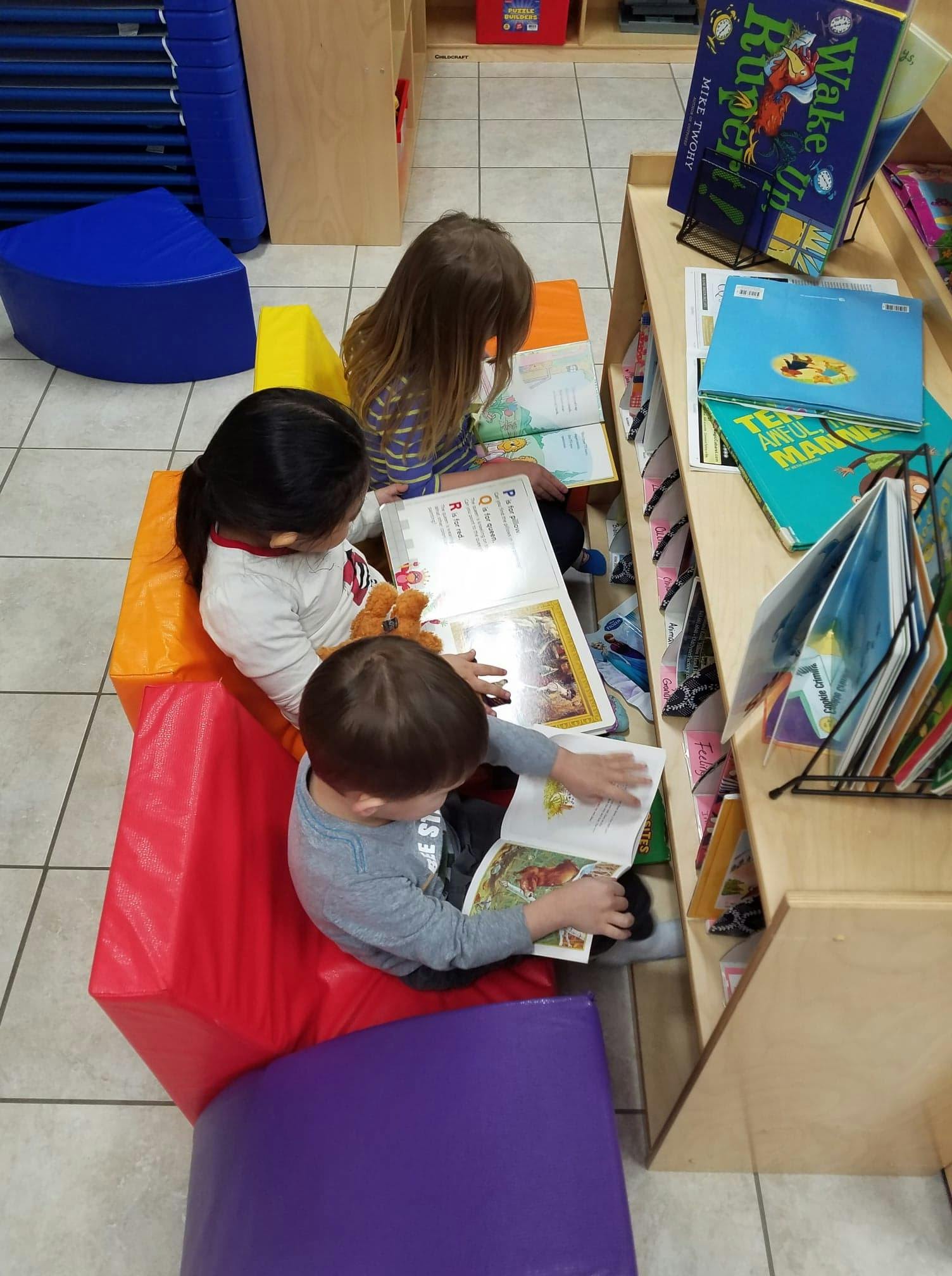 Three children sitting on colorful blocks reading books in a classroom