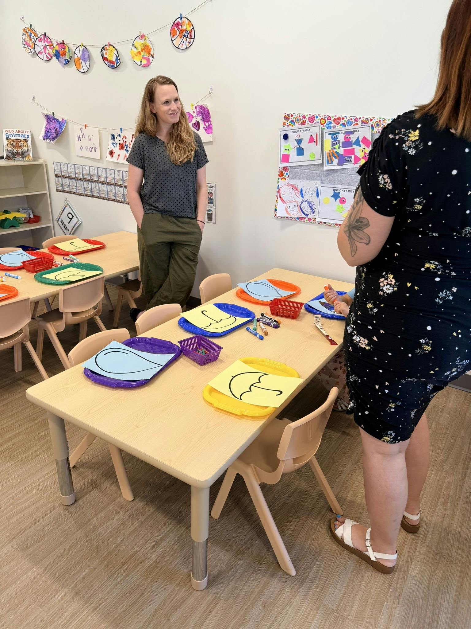 Two people standing and talking in a classroom next to low tables with crafts