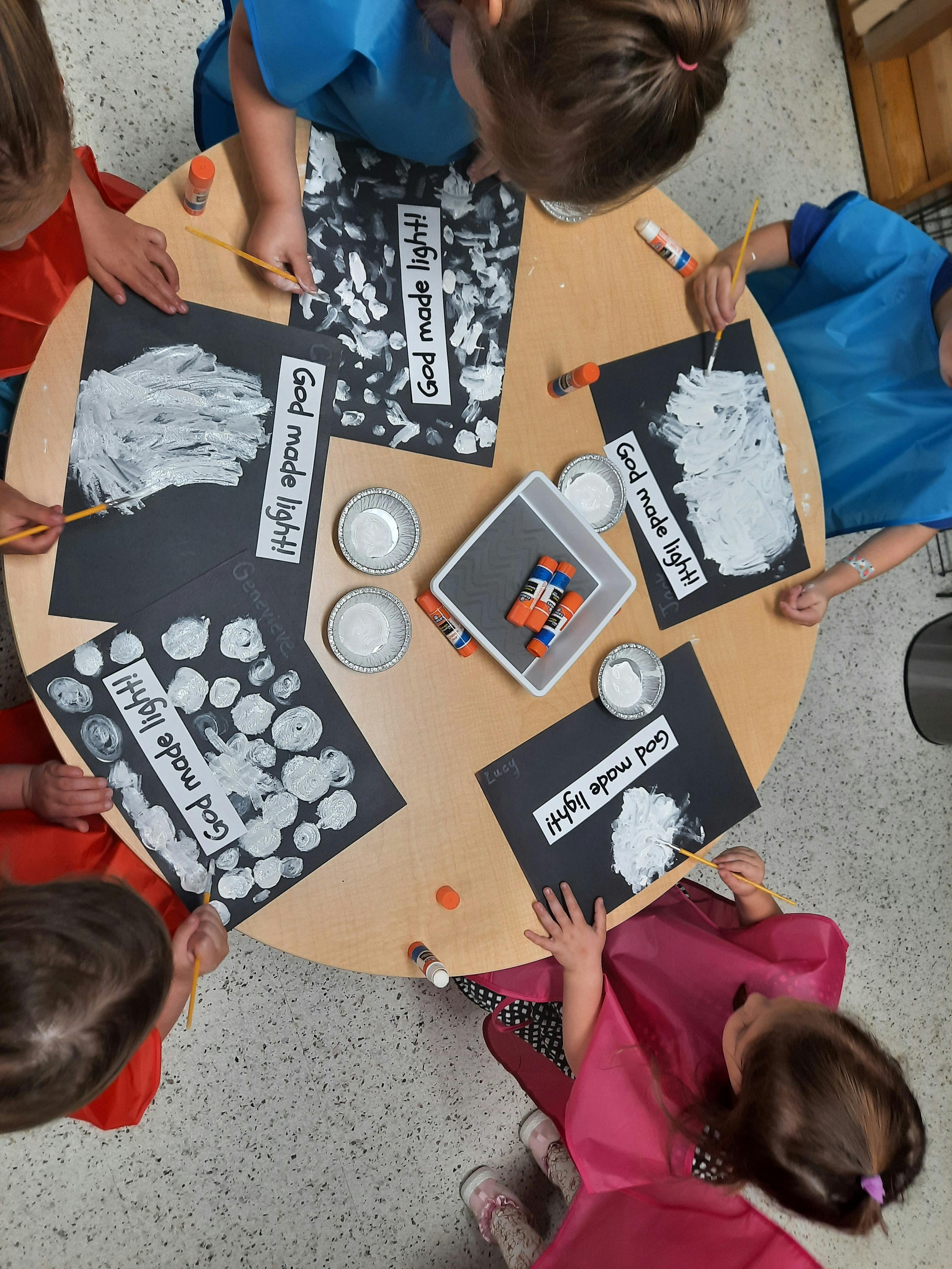 Children painting white shapes on black paper labeled God made light