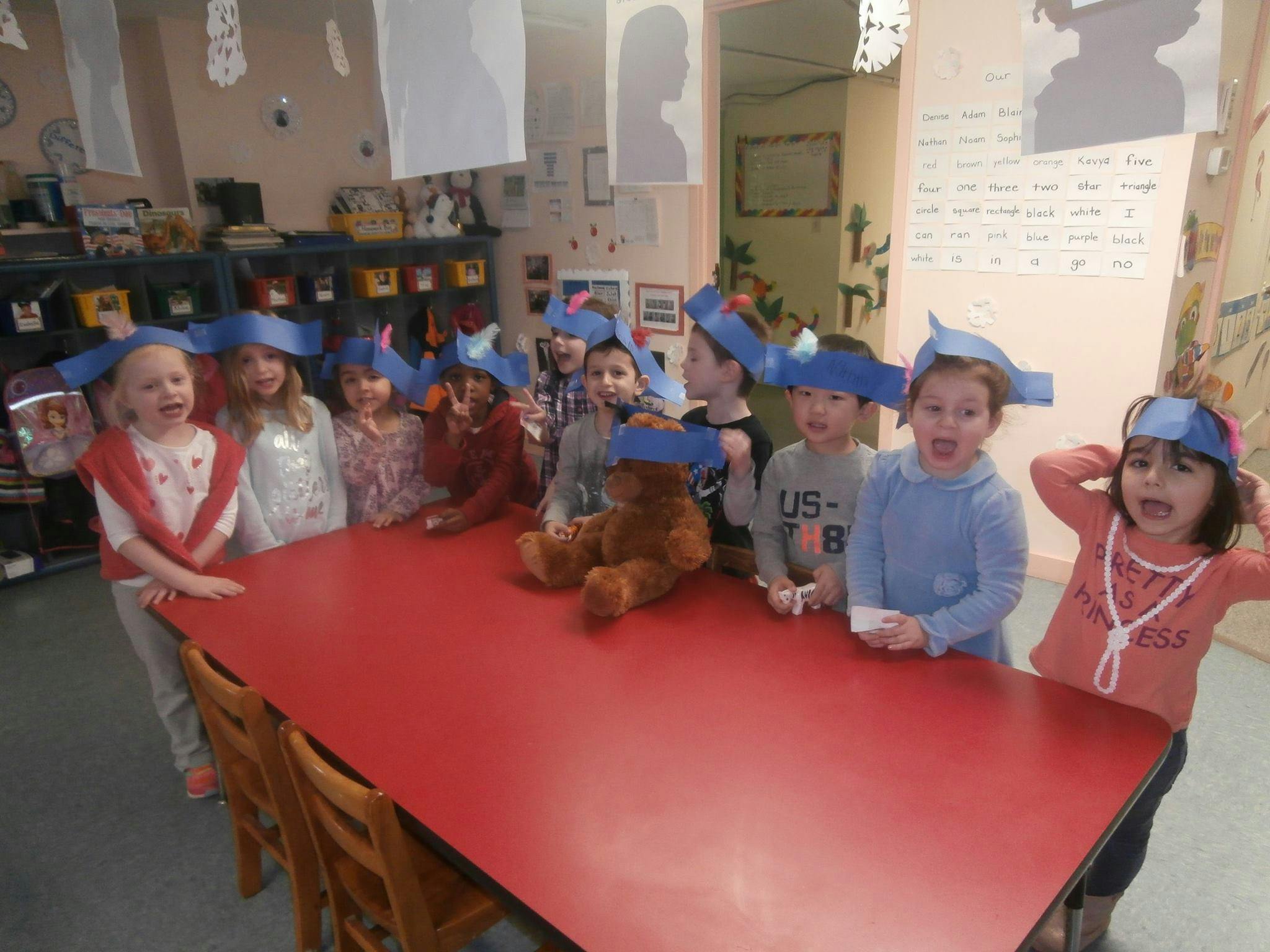 Children wearing blue paper hats standing around a red table with a teddy bear