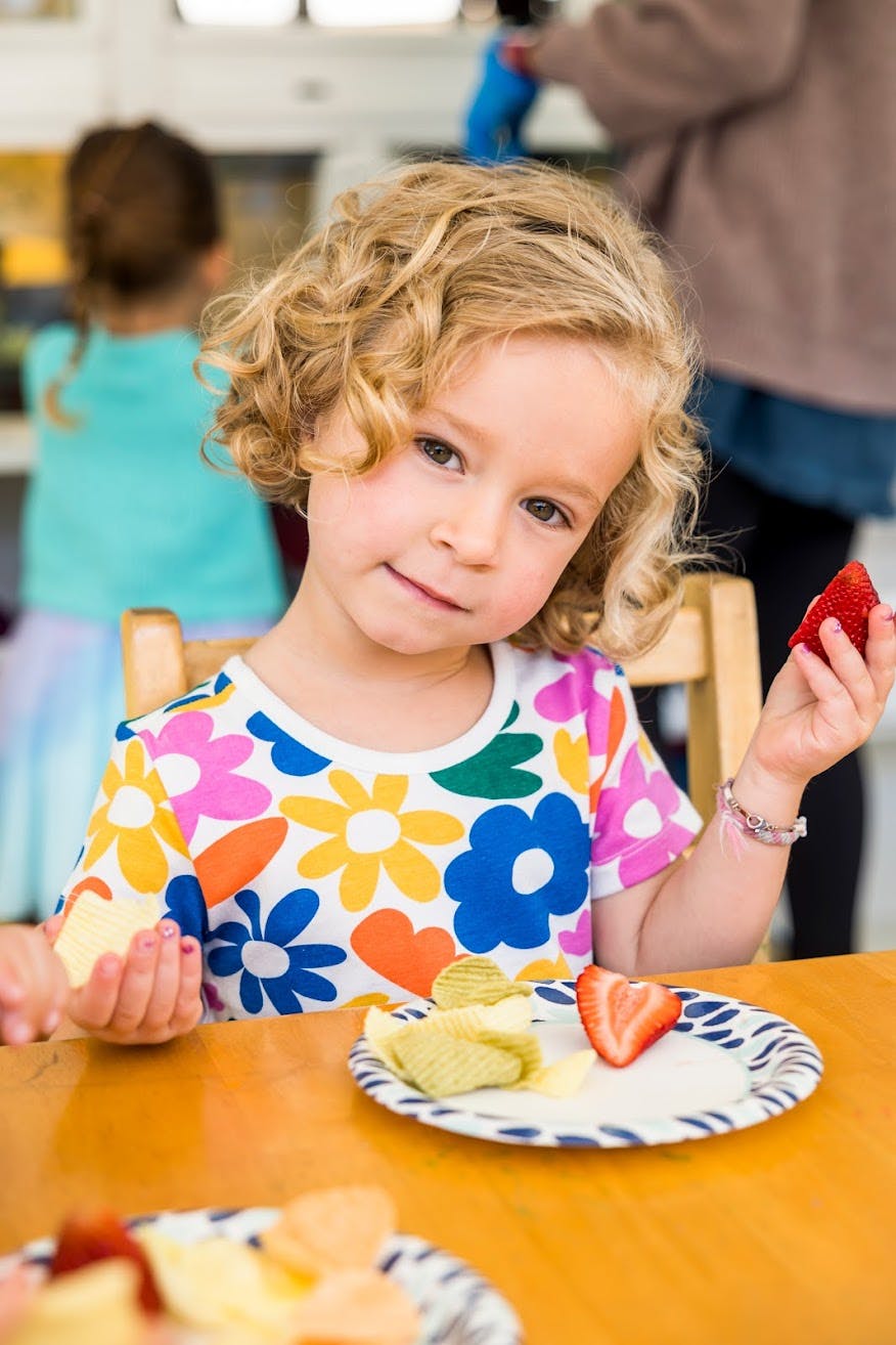 Child holding a strawberry and a piece of fruit, with a plate of snacks
