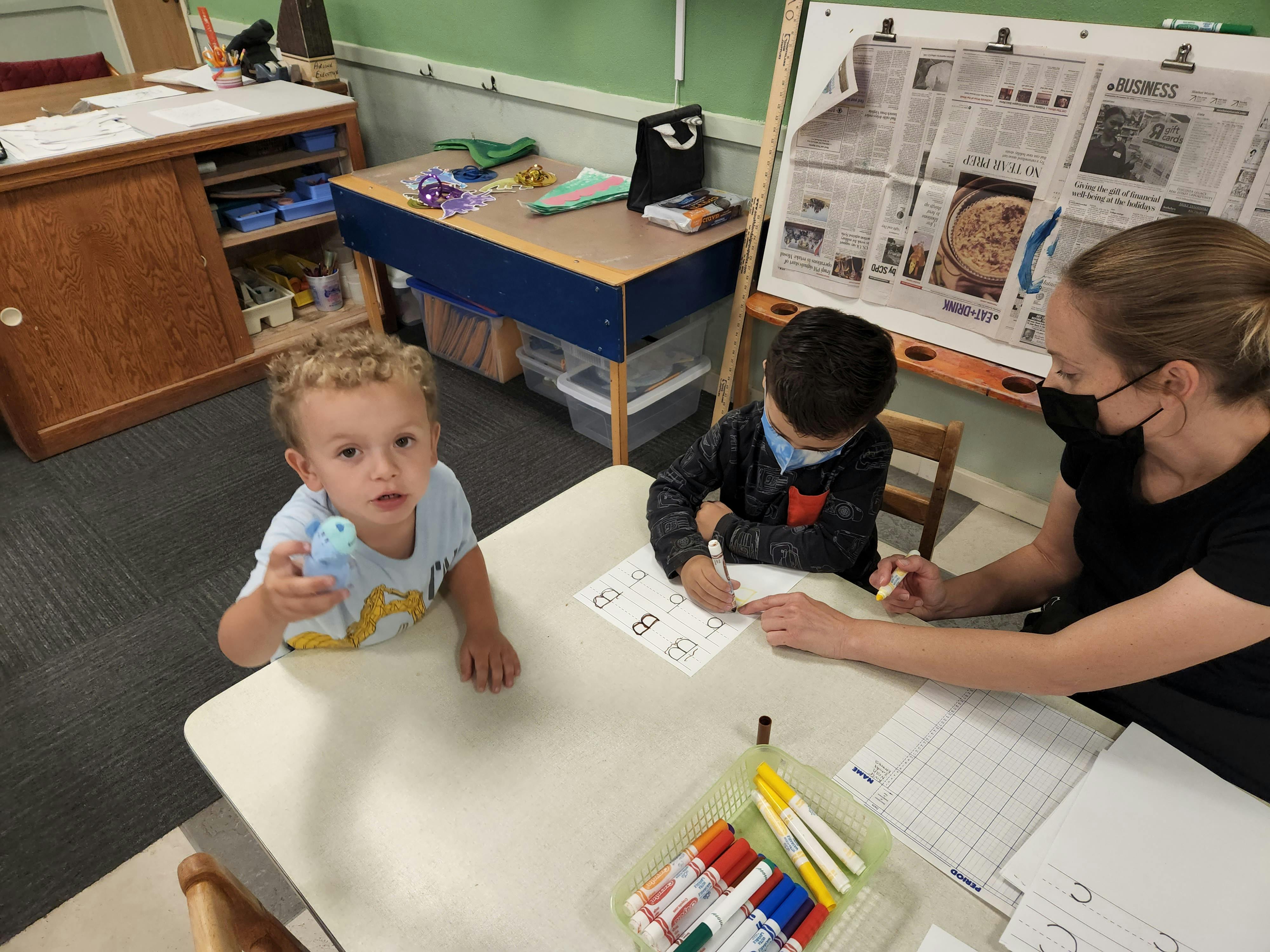 Adult and two children at a table, one child writing, another holding a toy