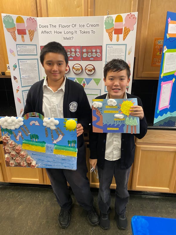 Two children in front of science fair projects, one holding a water cycle diorama