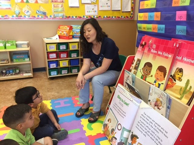 Adult sitting on a chair talking to two children sitting on a mat in a classroom
