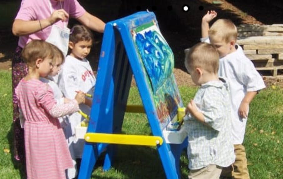 Adult and multiple children painting at an easel outdoors
