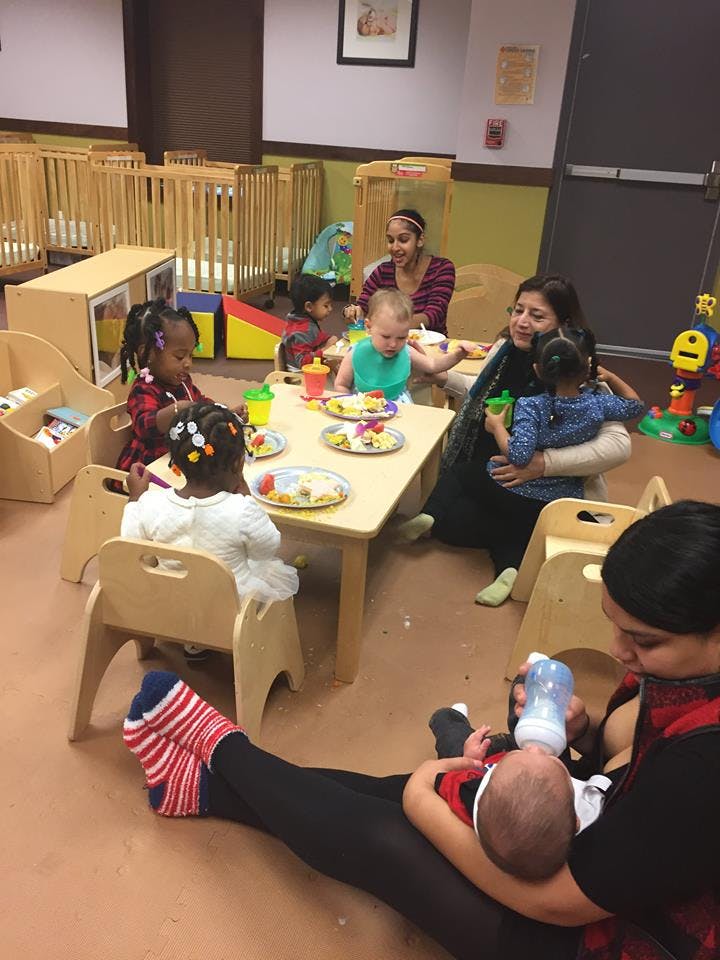 People and children in a classroom, some eating at a table, one person feeding an infant