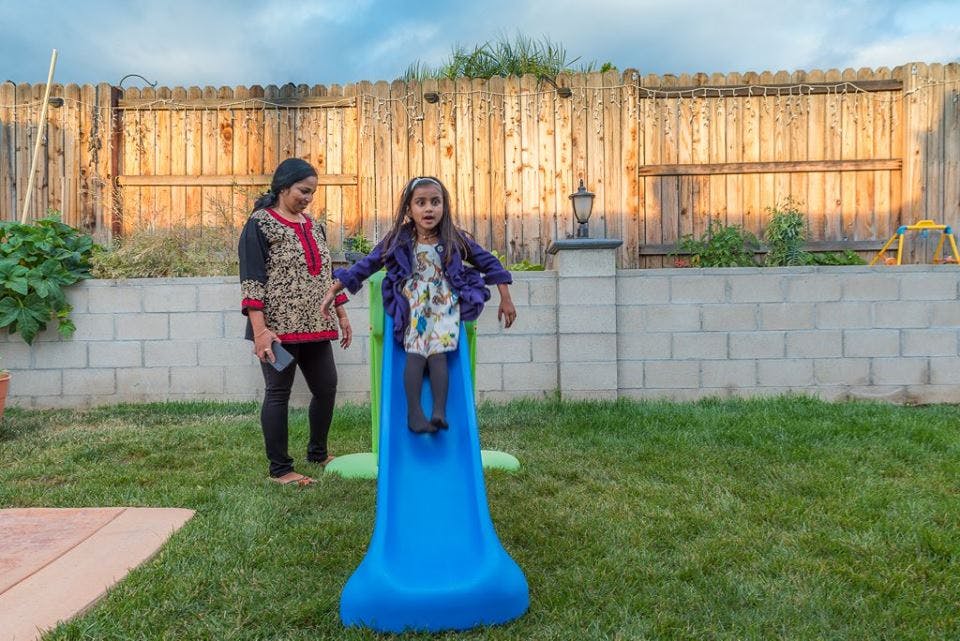 Person standing next to a child on a blue slide in a grassy yard