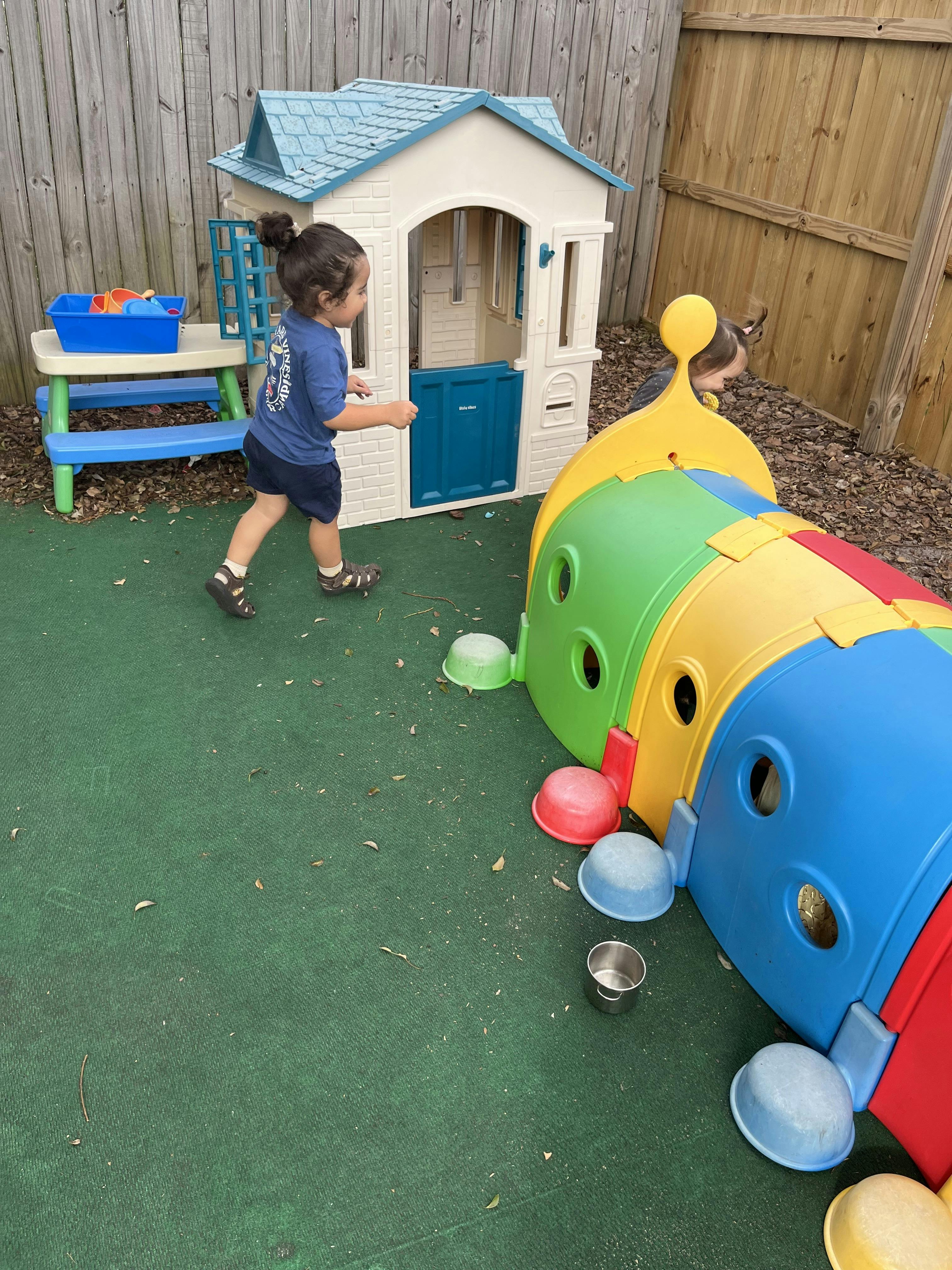 Child running past a playhouse and a colorful tunnel on green turf