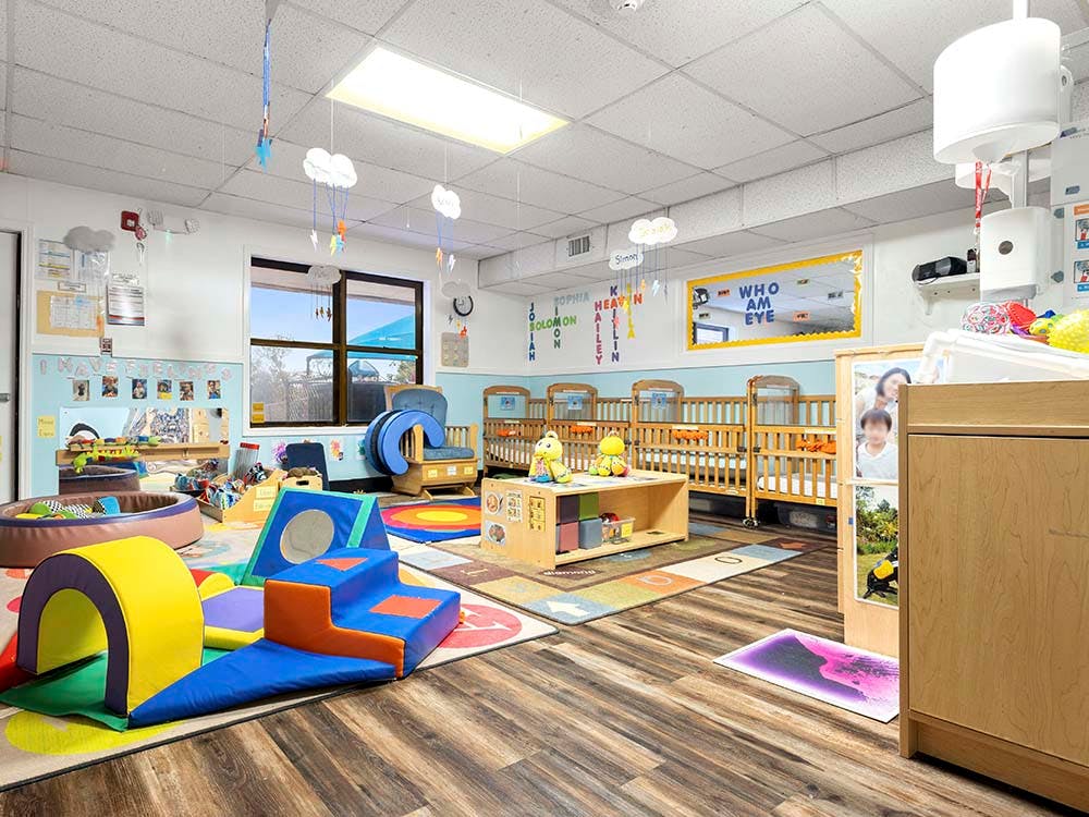 A childcare classroom with cribs, play mats, and a window