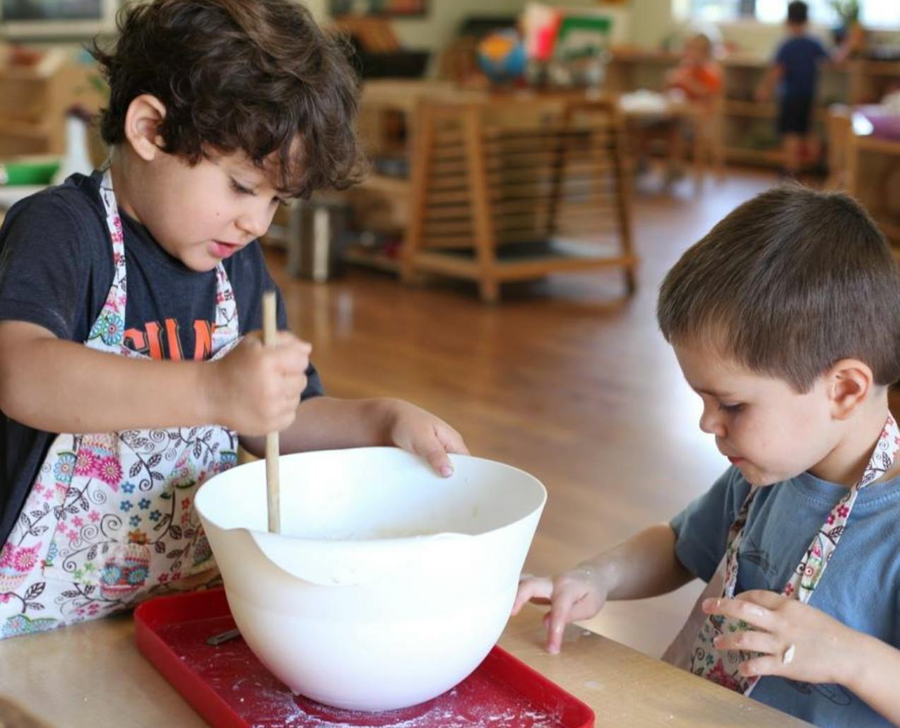 Child stirring a bowl with a wooden spoon, another child watching