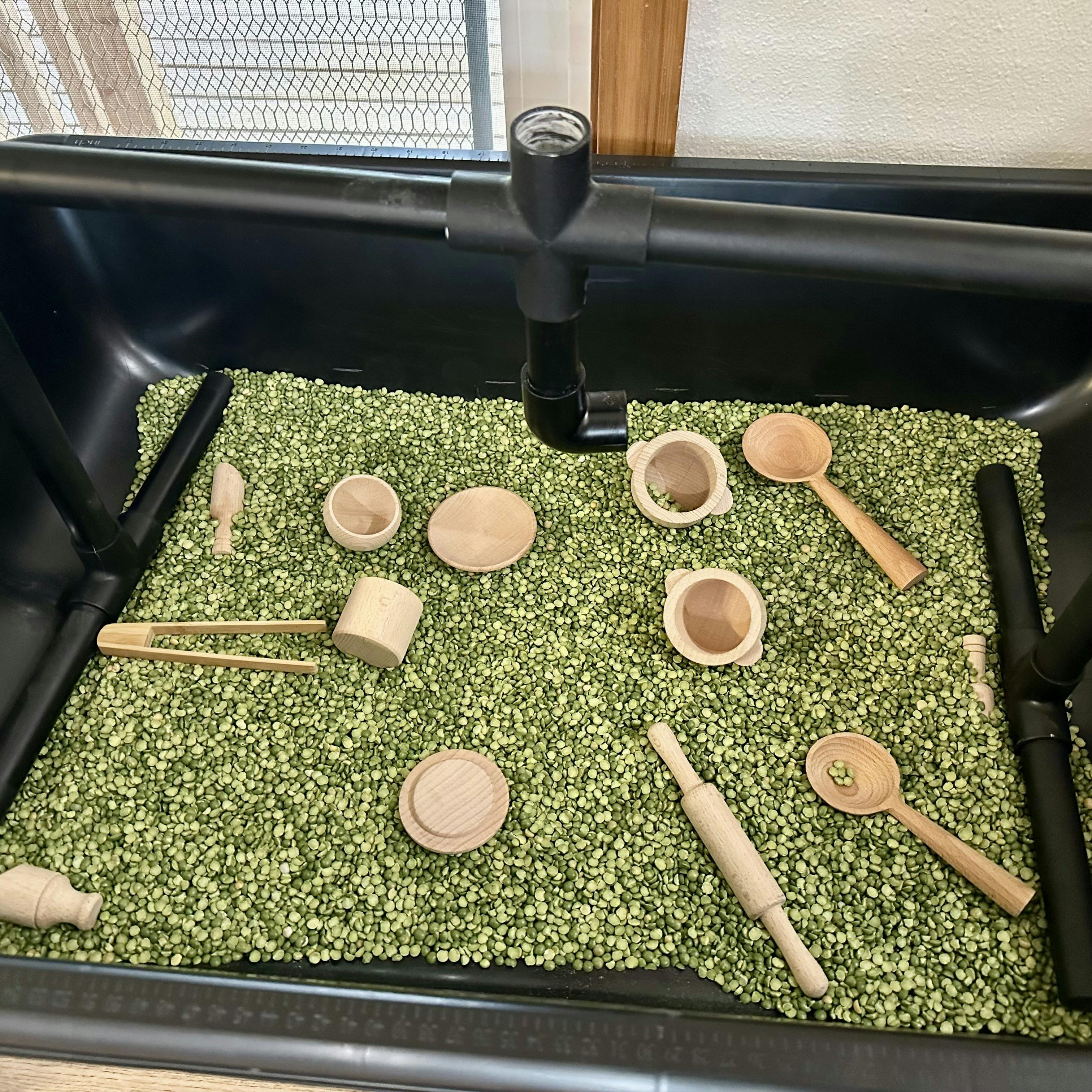 Wooden kitchen play items on a bed of green split peas in a black container