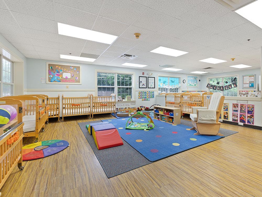 A brightly lit nursery room with multiple cribs, a blue play mat, and toys