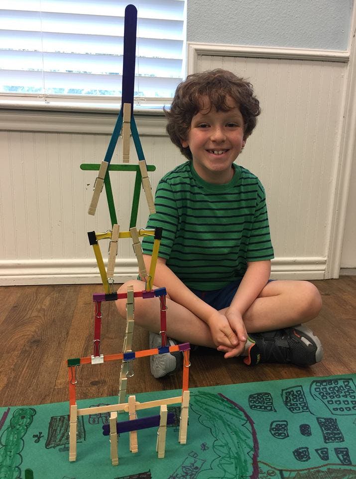 Child sitting next to a tall structure made of craft sticks and clothespins