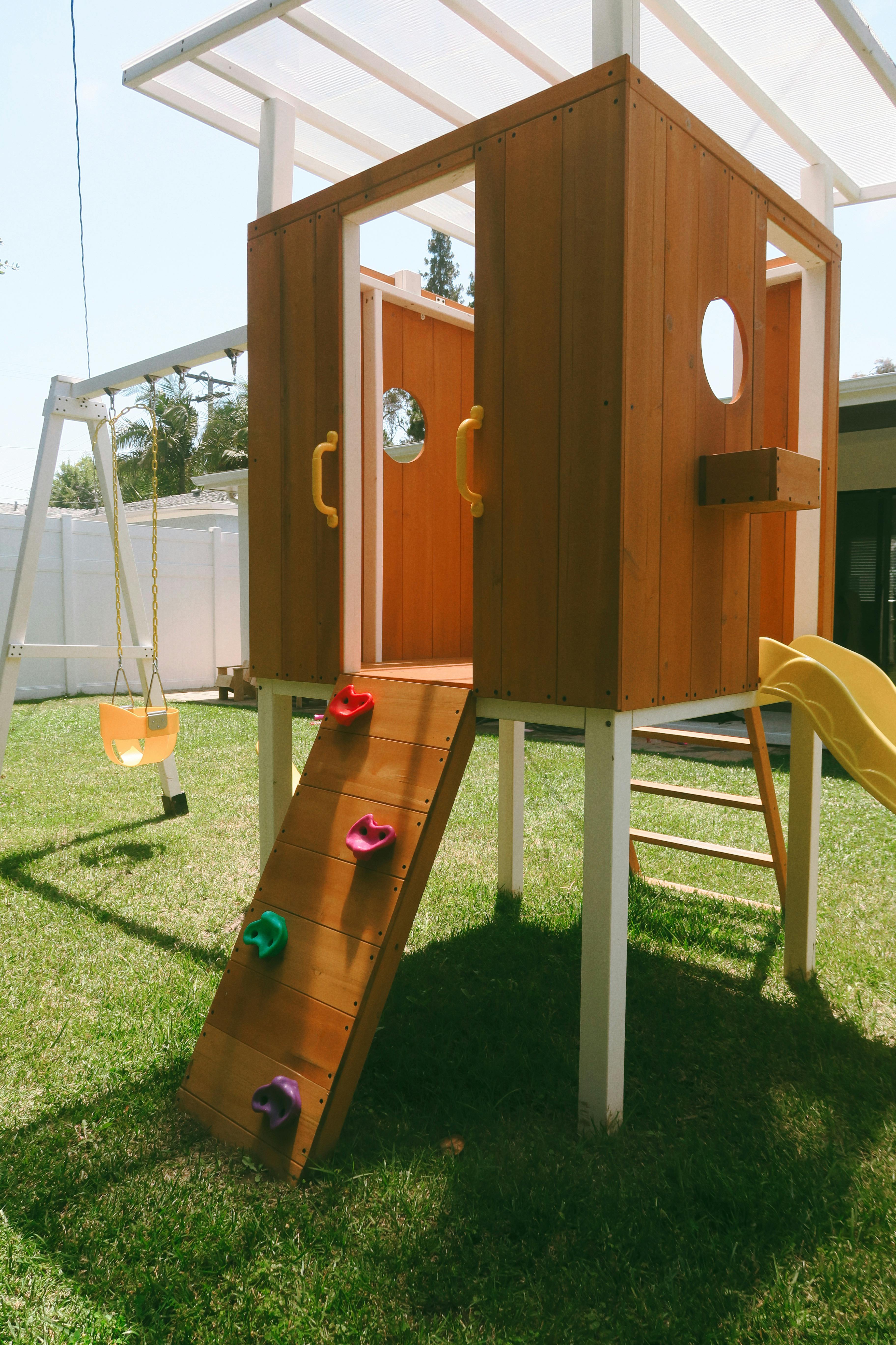 Wooden playhouse with a climbing wall, slide, and swing set