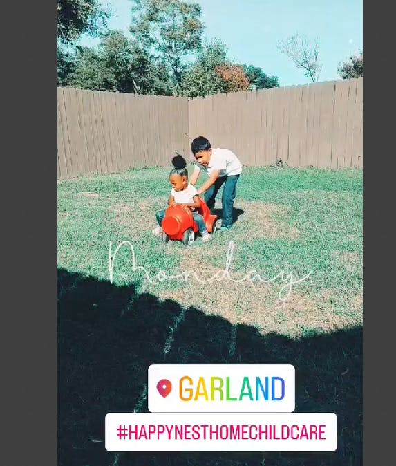 Child sitting in a toy car with another child pushing it in a grassy yard