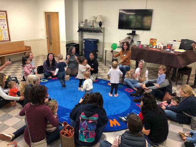 Adults and children sitting and standing on a blue rug in a room