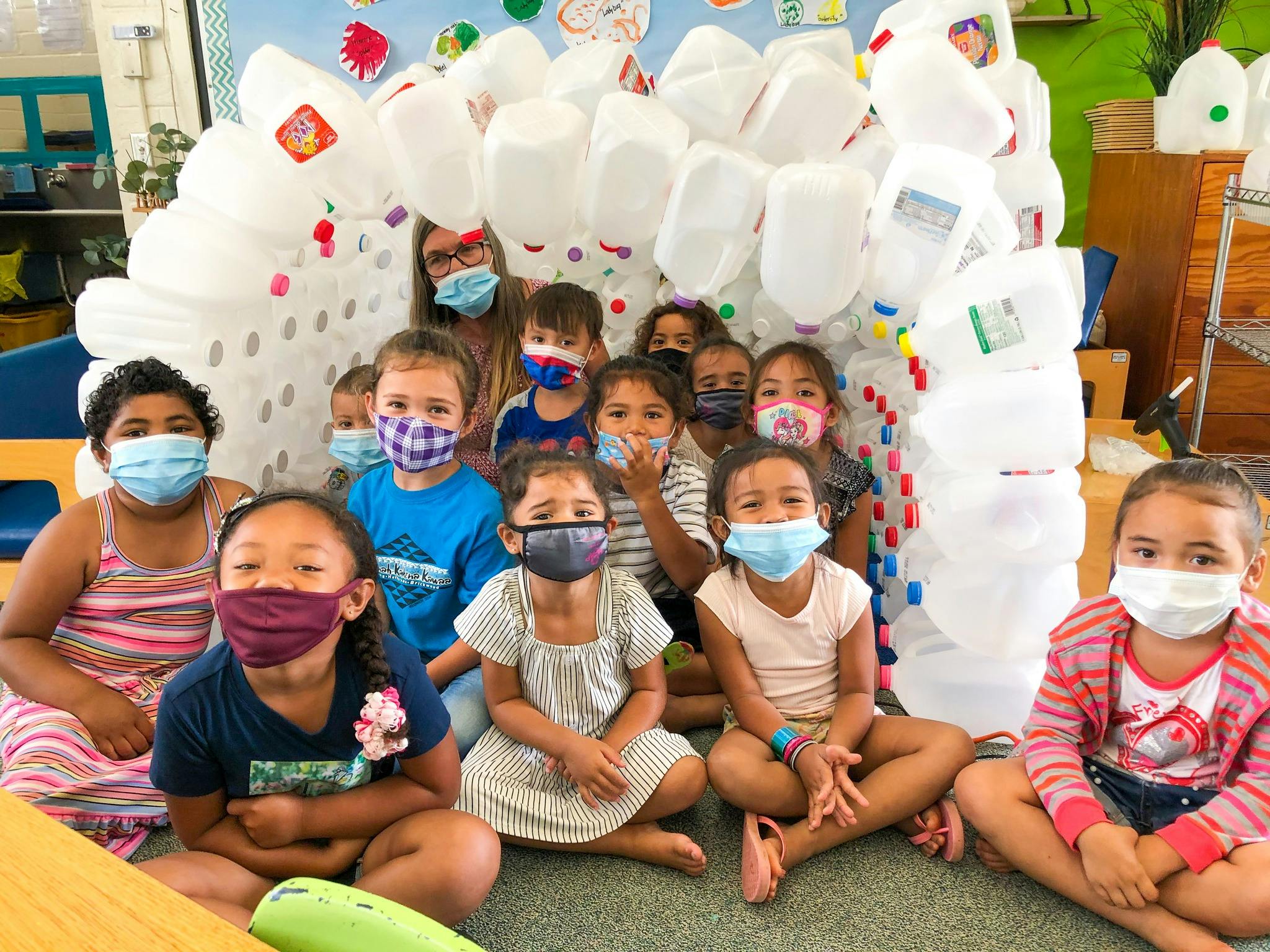 Group of children and one adult wearing masks, sitting in front of a structure made of plastic jugs