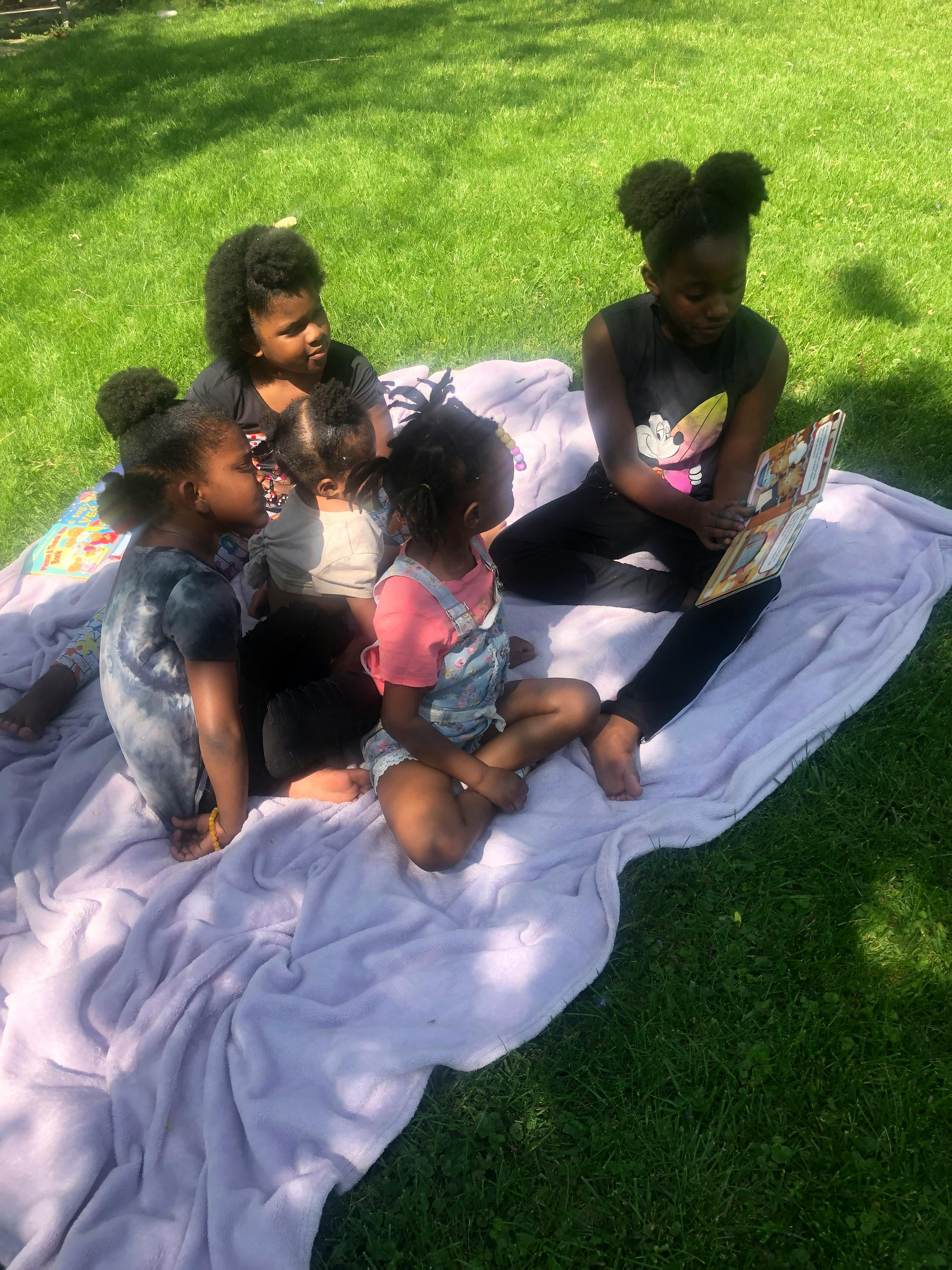 Children sitting on a blanket on grass, one child reading a book