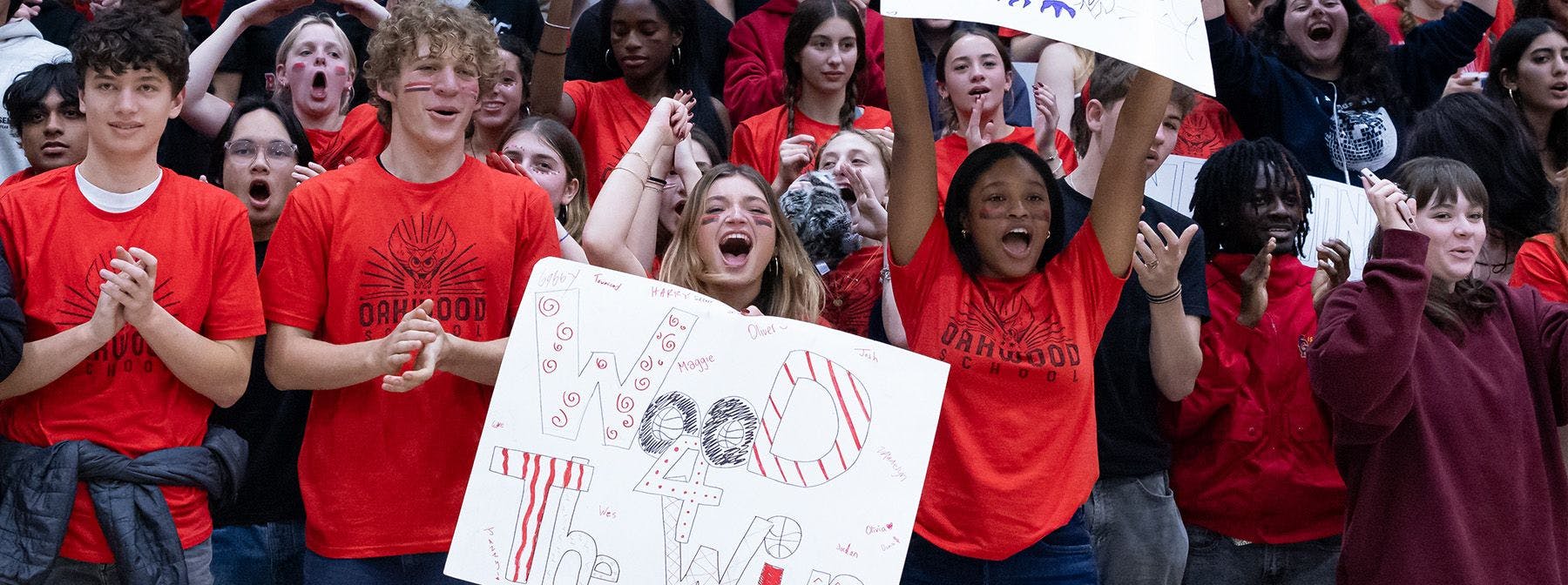 Group of cheering teens in red shirts, one holding a sign that says "OAKWOOD SCHOOL"