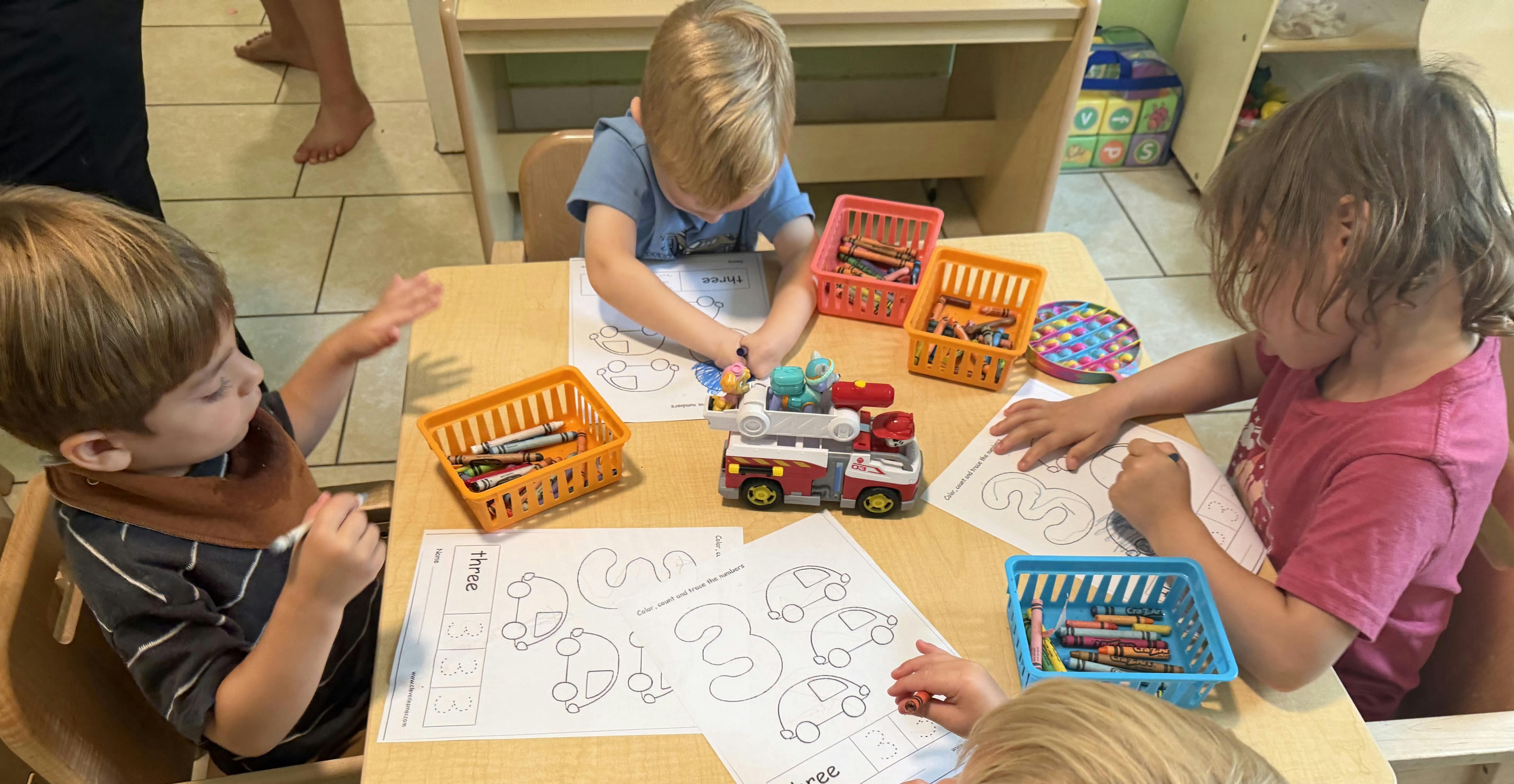 Children sitting at a table coloring worksheets and playing with a toy fire truck