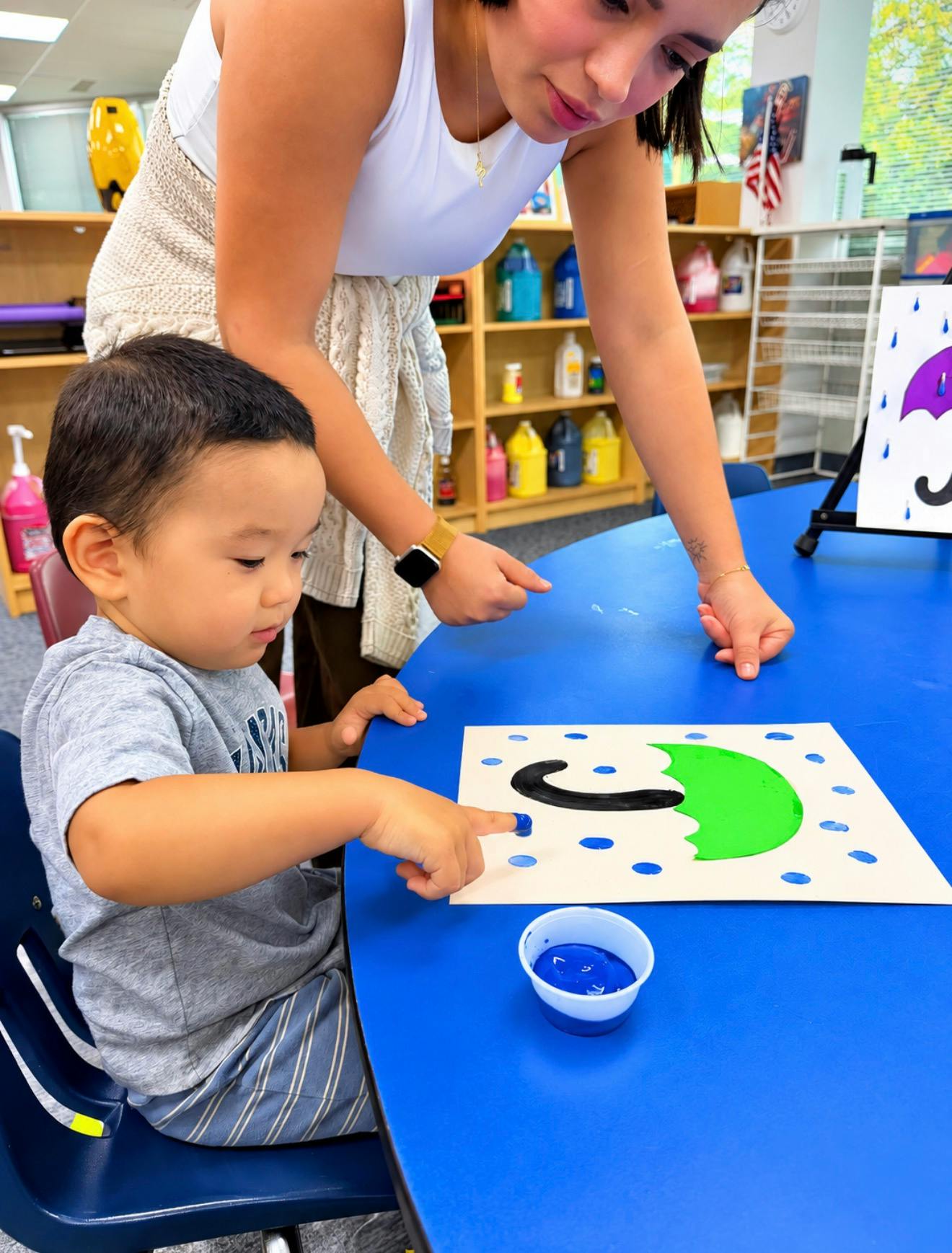 A person helping a child paint raindrops on an umbrella craft