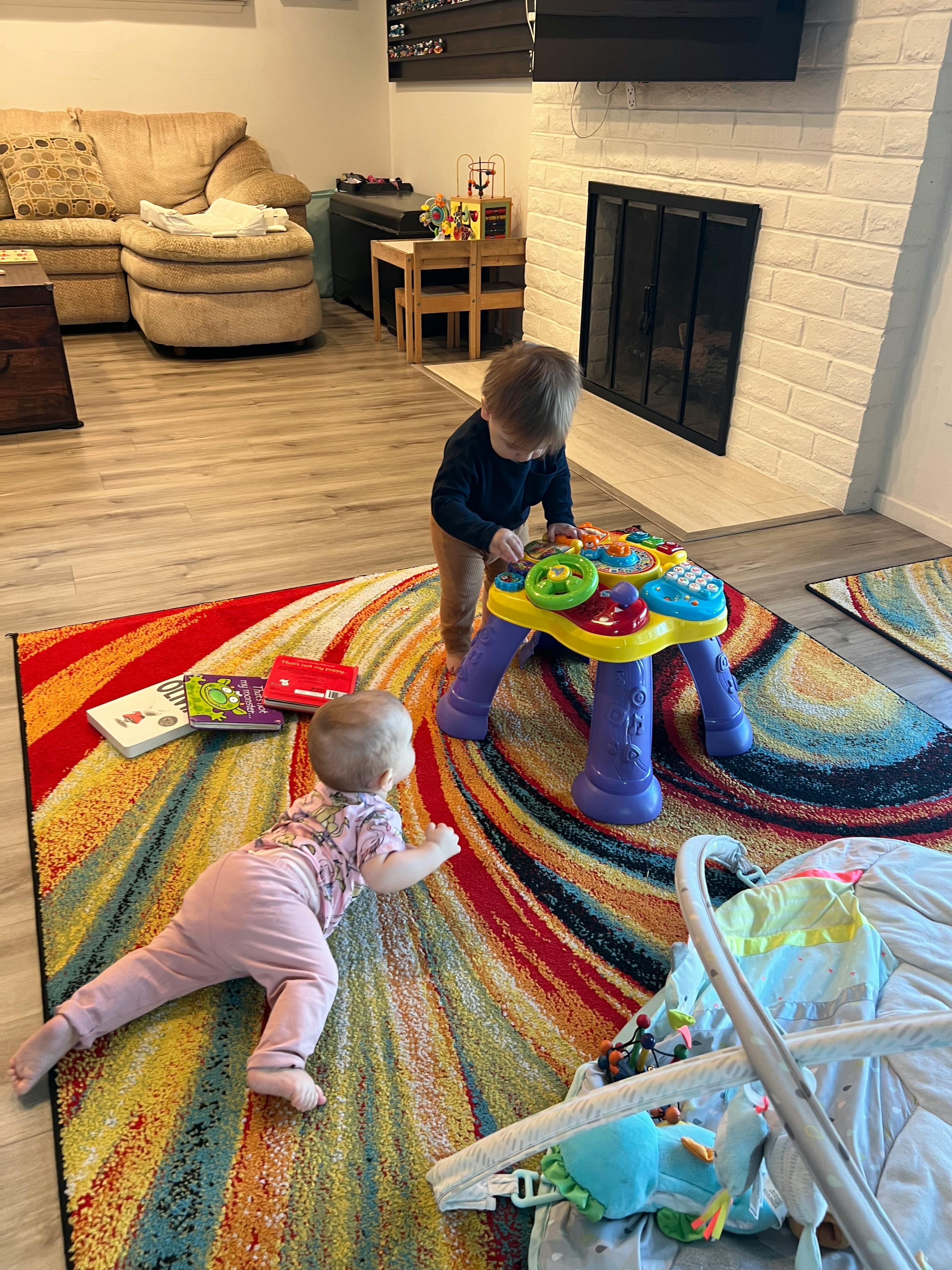 Child standing at a toy table, another child crawling on a rug