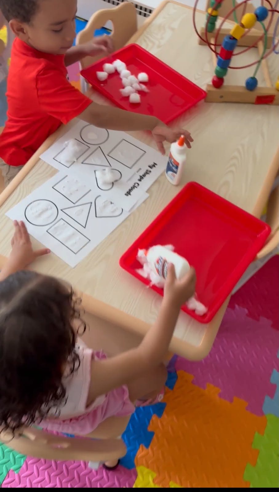 Children crafting with cotton balls and glue on a shape activity sheet