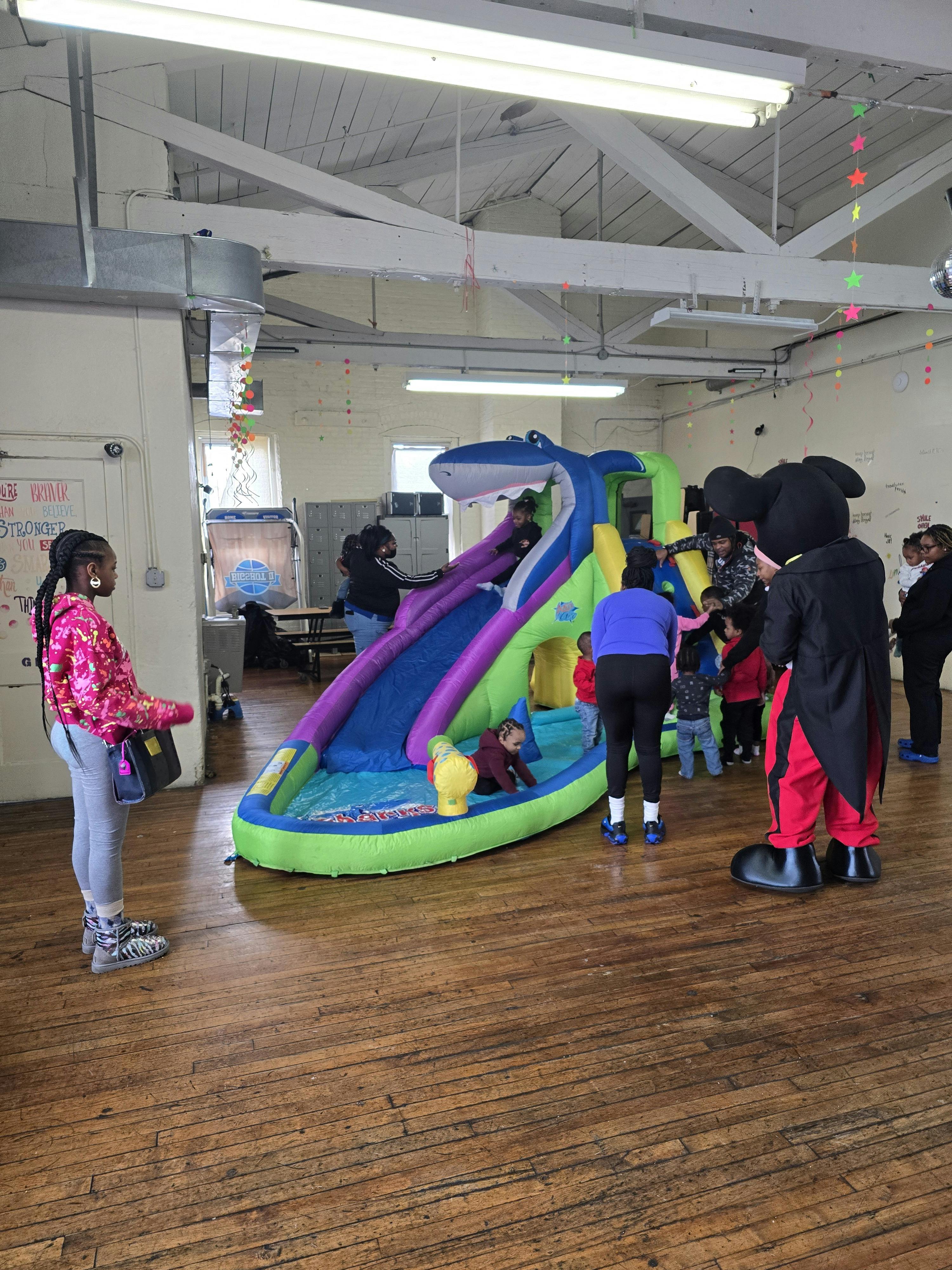 People and a person in a mascot costume at a party with a shark inflatable slide