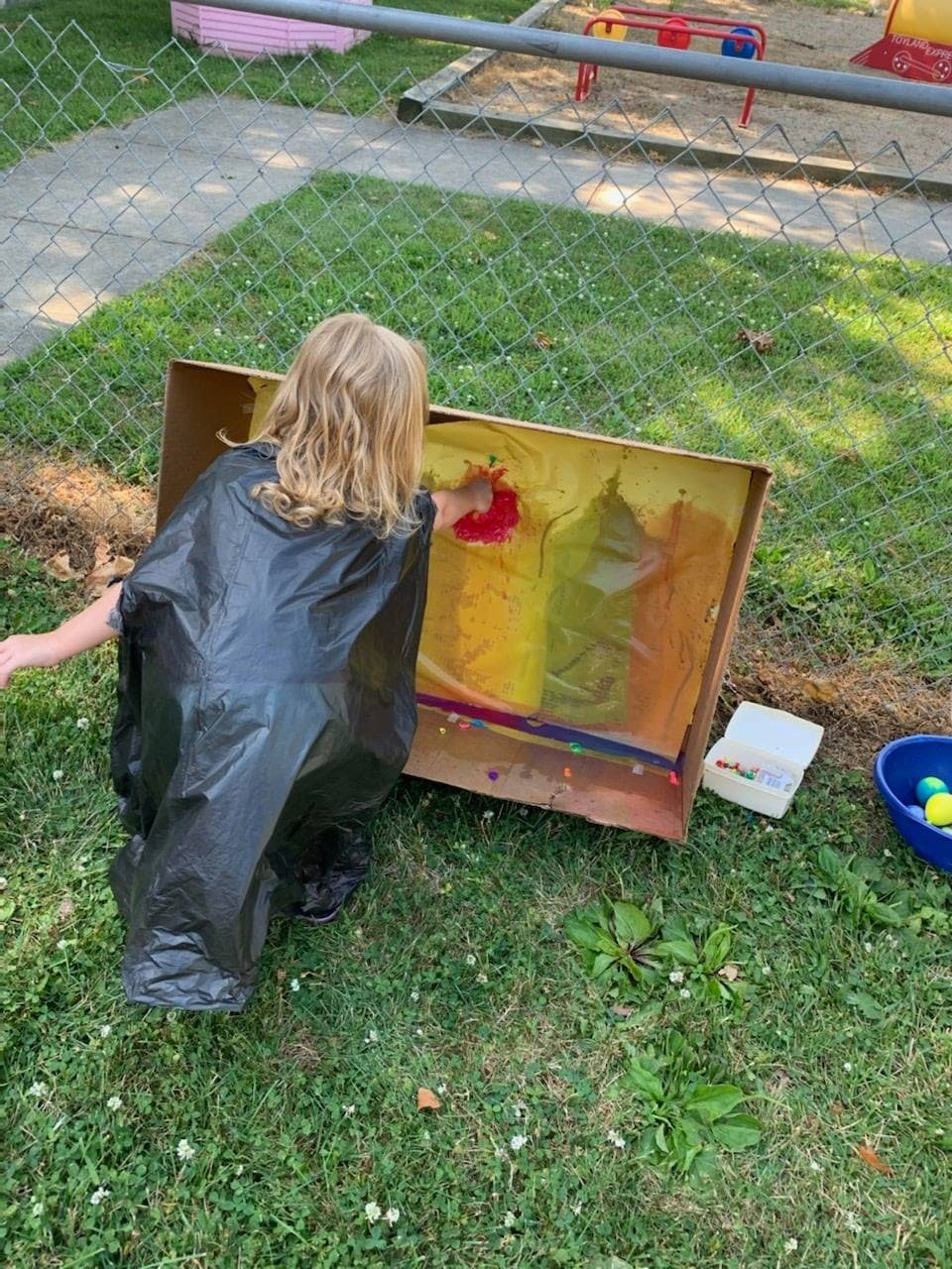 Child in a black smock painting on a cardboard box outdoors