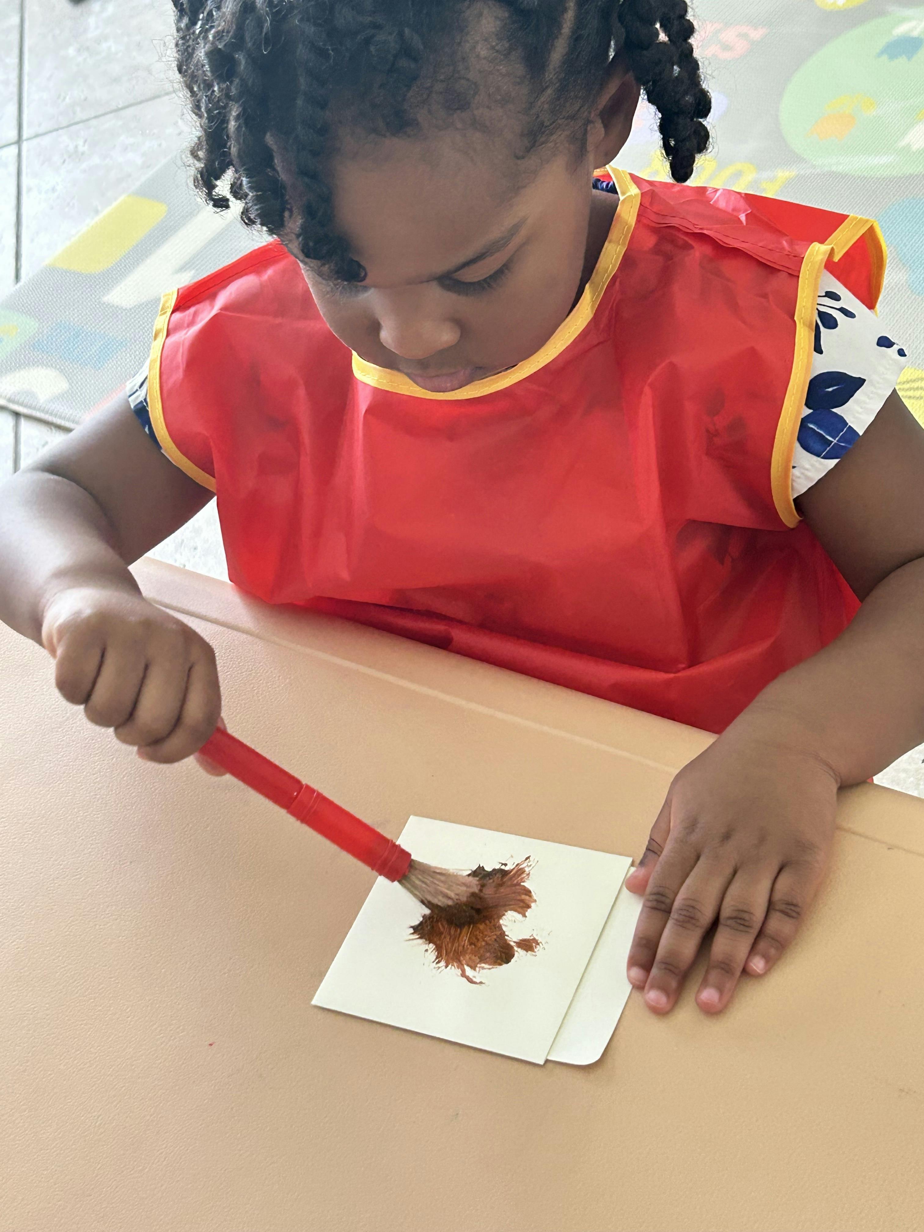 Child in a red apron painting on paper with a paintbrush
