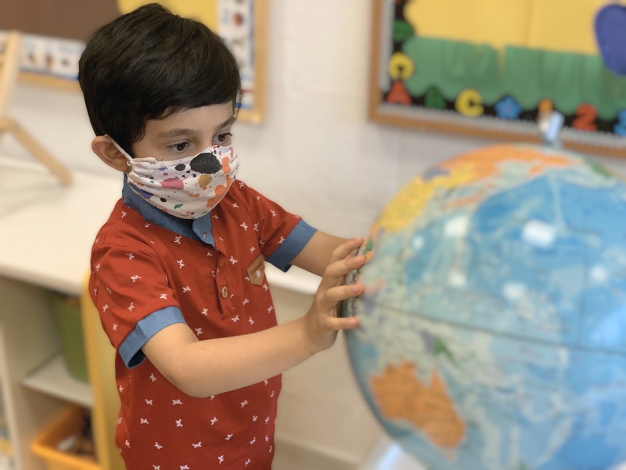 Child in a mask touching a globe in a classroom
