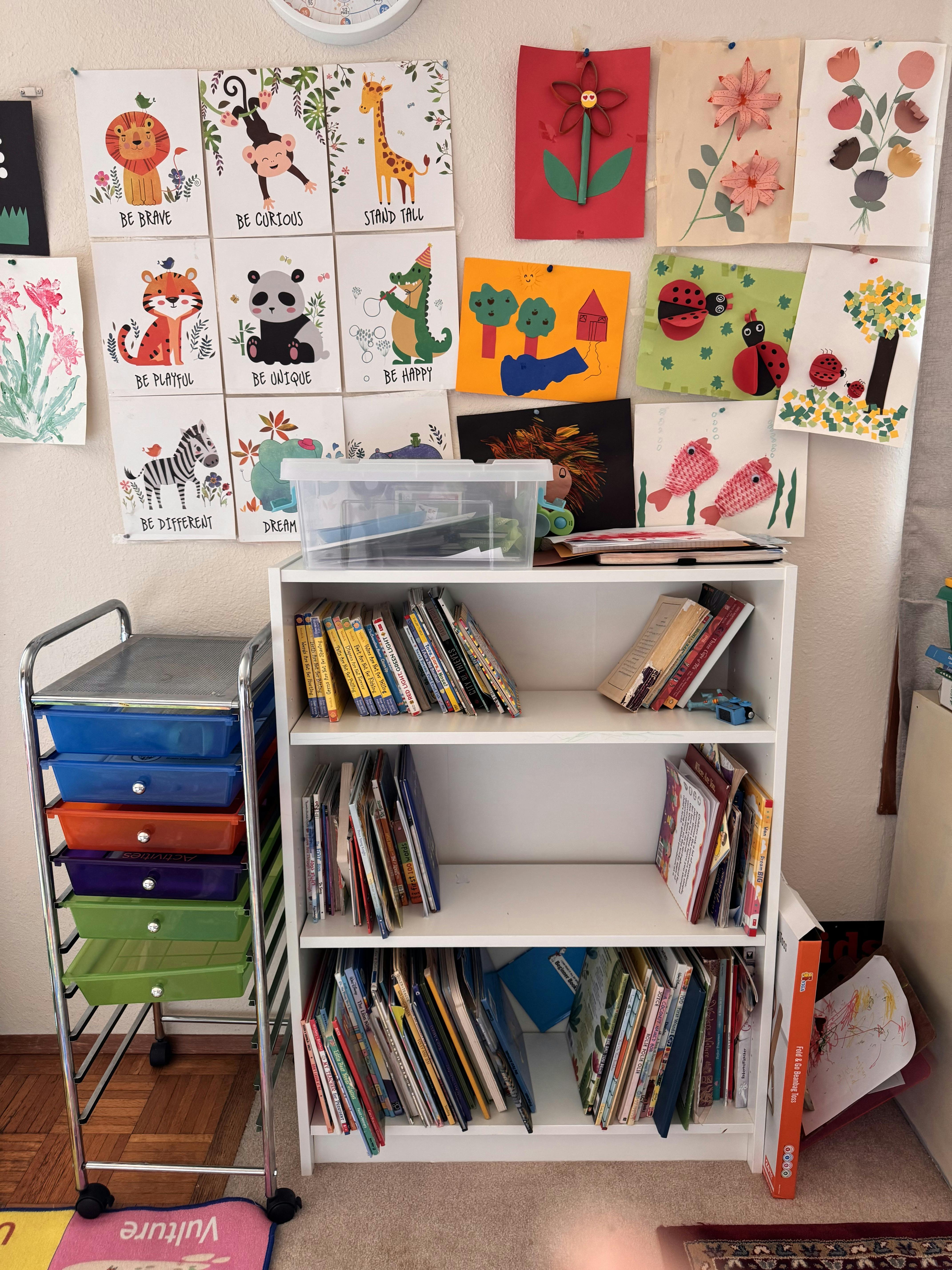 Children's artwork and books on shelves next to a rolling cart with colorful drawers