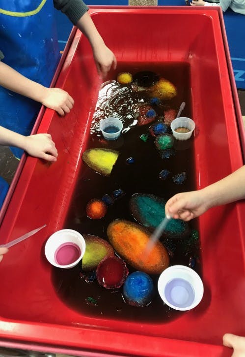 Children's hands playing with colorful objects in a red sensory bin with liquid