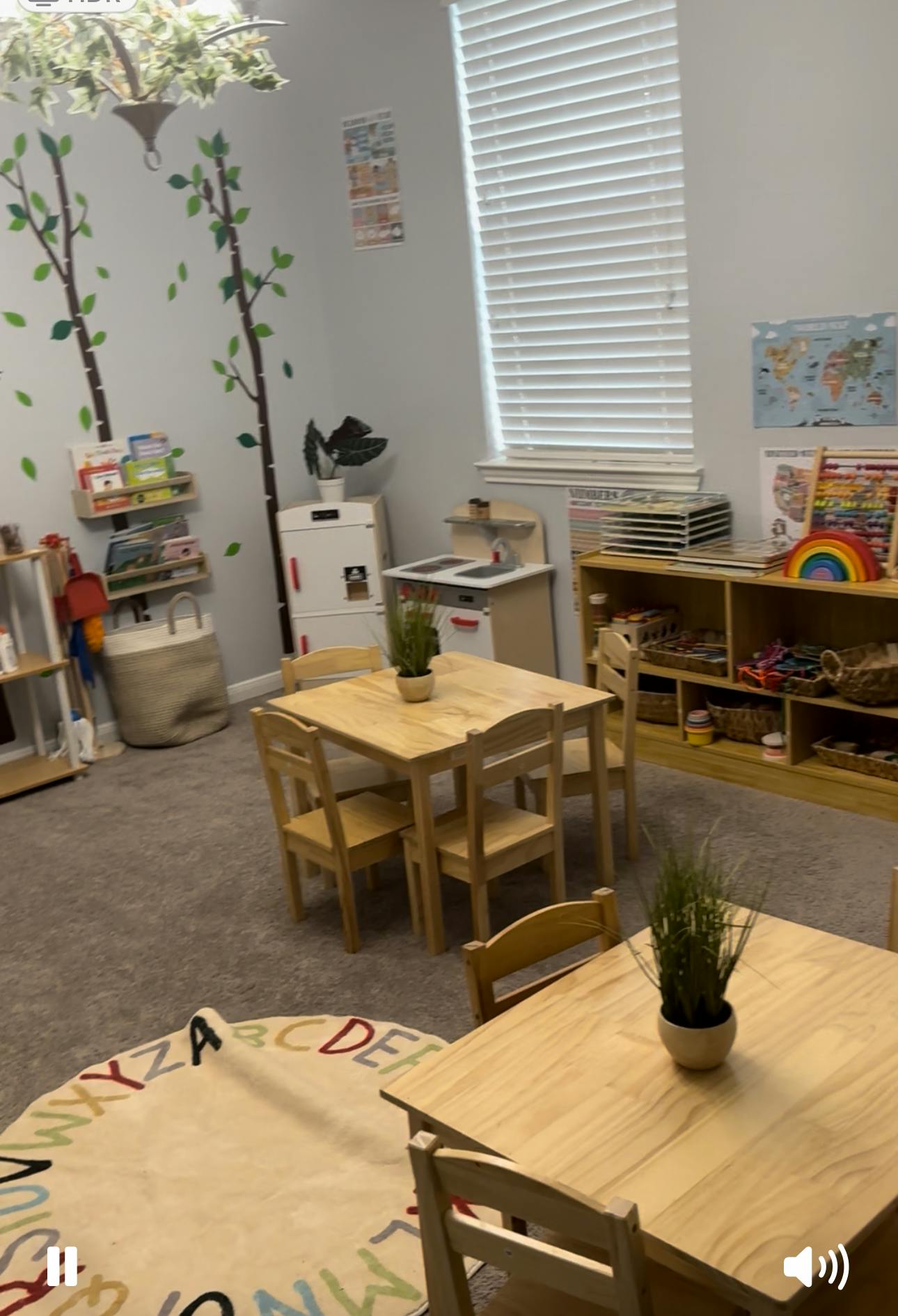 Classroom with small wooden tables and chairs, shelves with toys, and a window