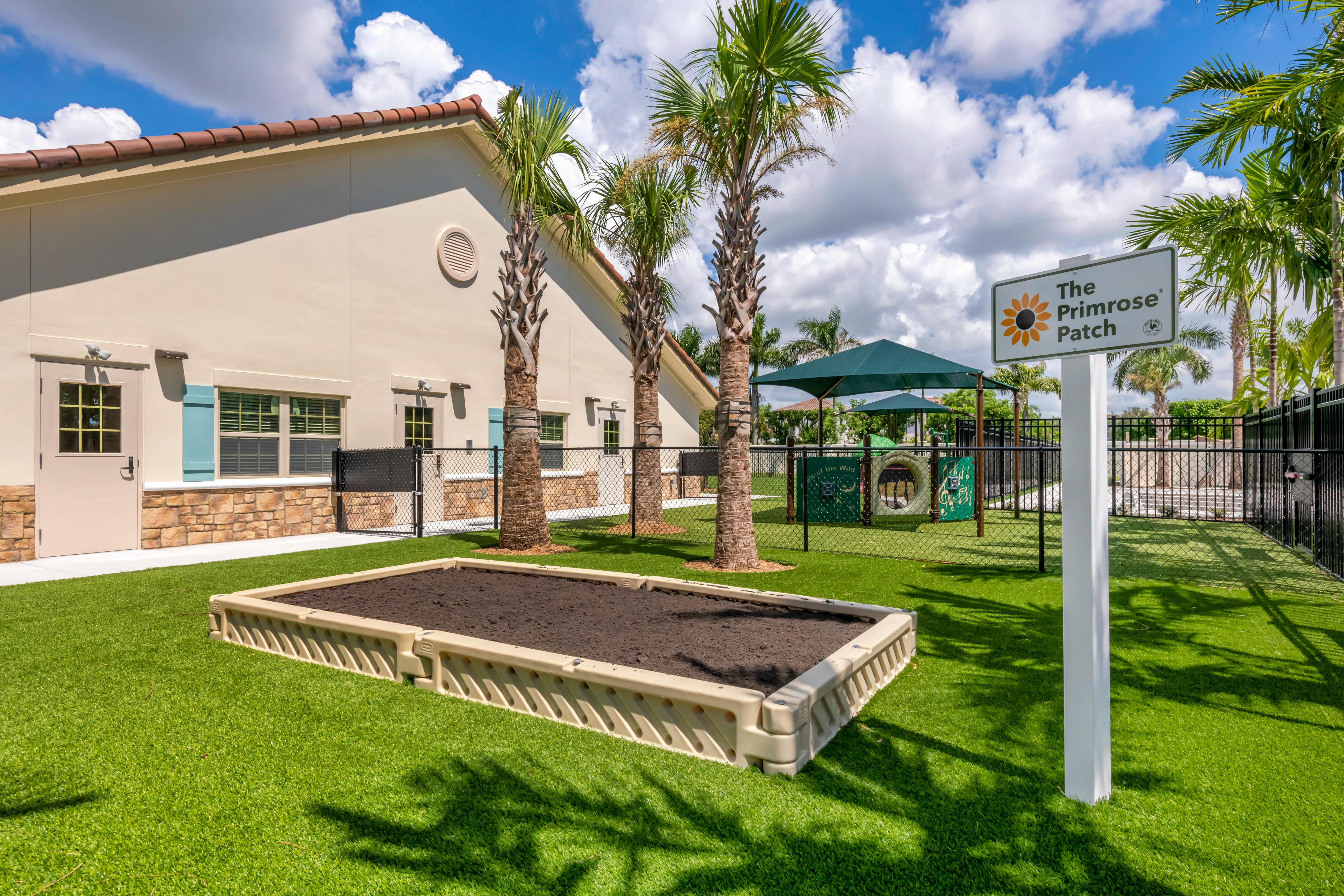 Raised garden bed filled with soil on artificial grass next to a building and palm trees