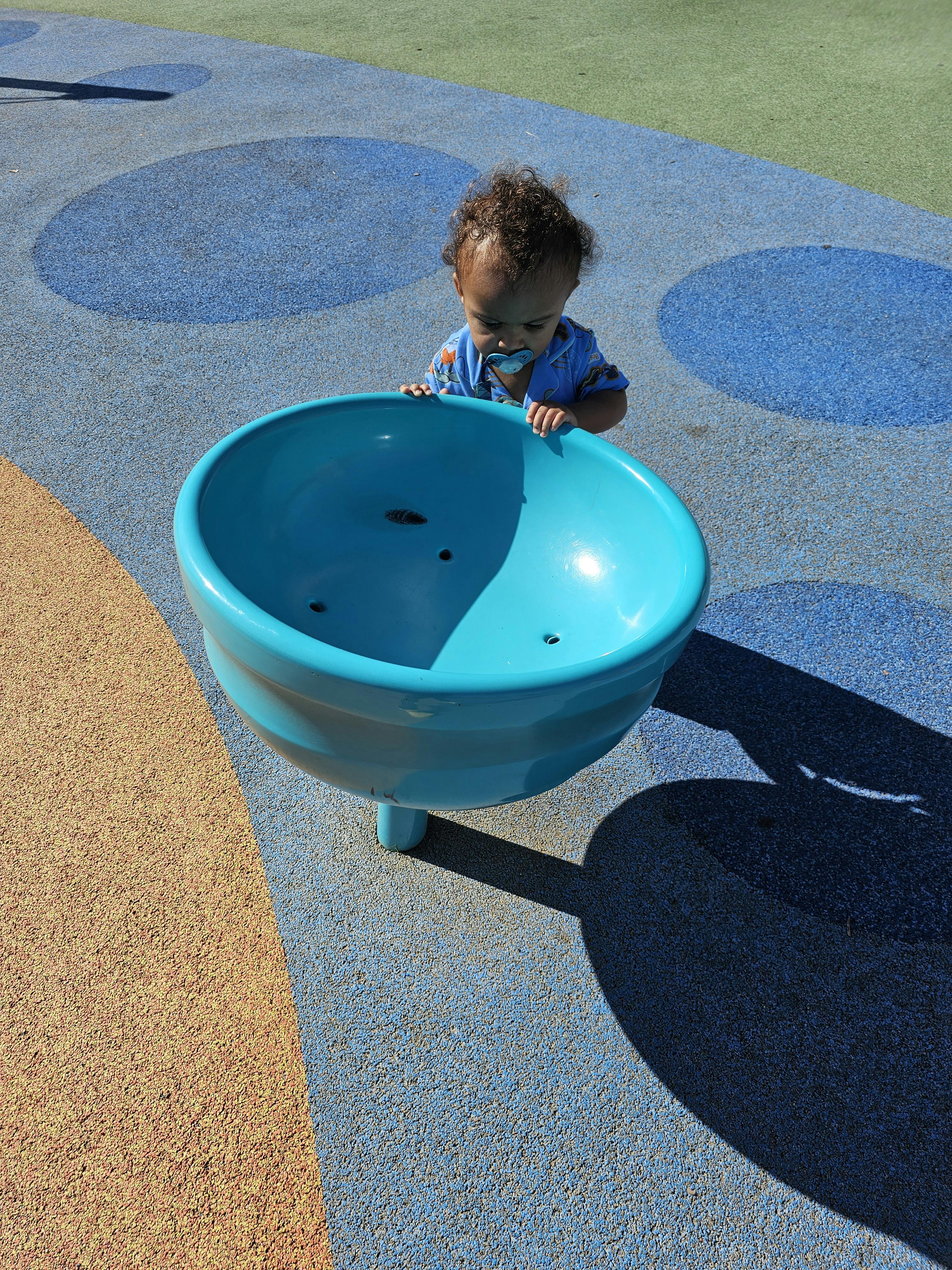 Child with a pacifier looking into a blue bowl on a playground