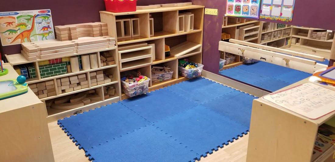 Classroom with wooden shelves, blue floor mats, and a dinosaur poster