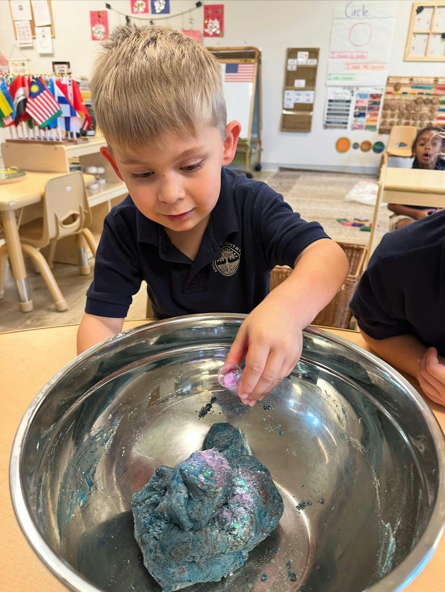 Child adding glitter to blue dough in a metal bowl in a classroom