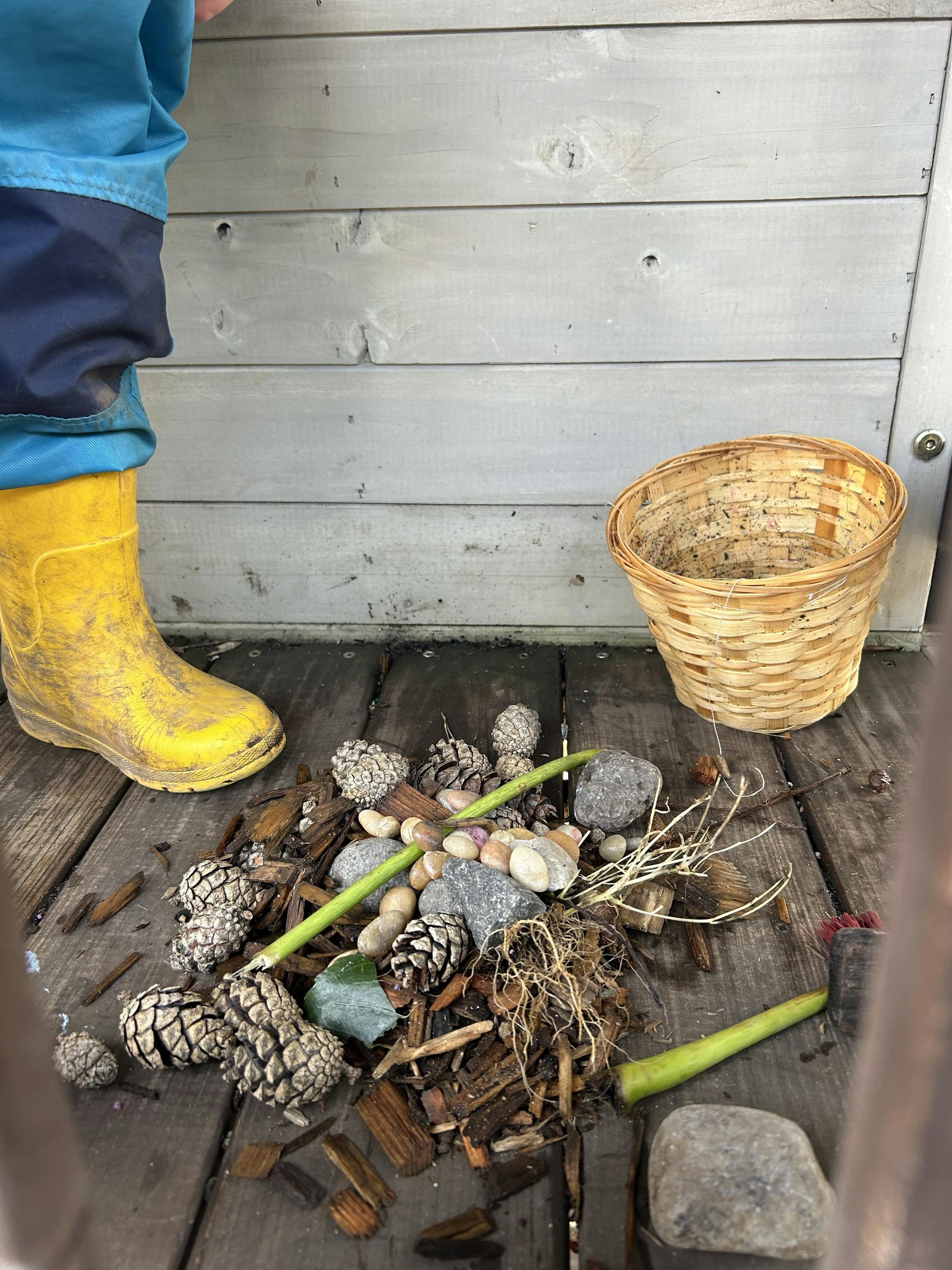 Child in yellow boot next to a pile of pinecones, sticks, rocks, and roots