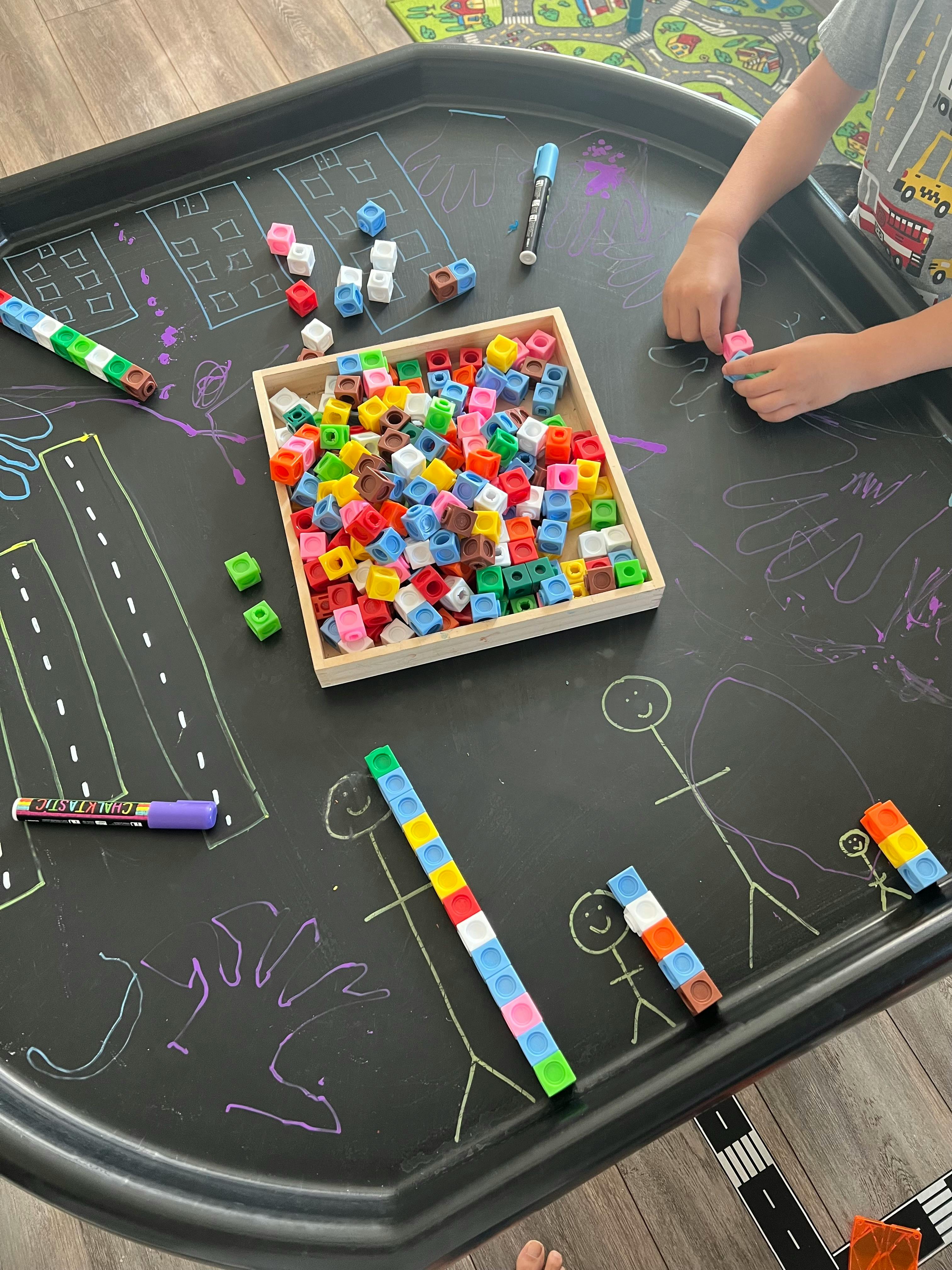 Child playing with colorful connecting cubes on a black tray with chalk drawings