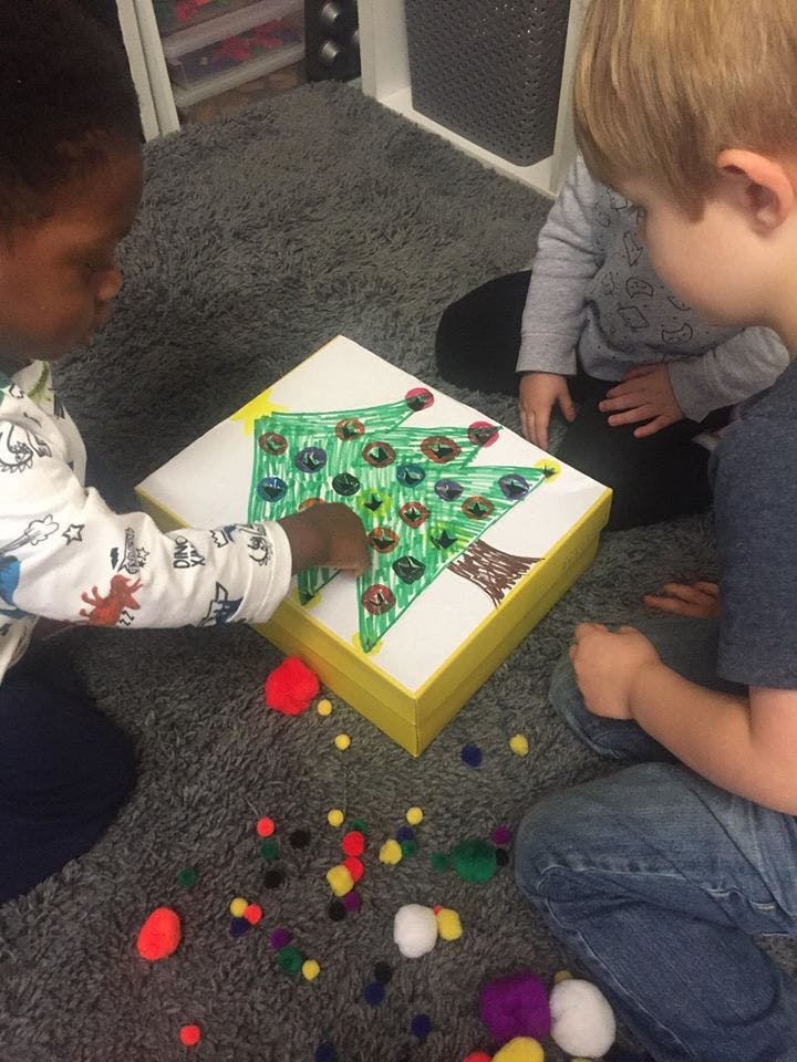 Two children decorating a box with a drawn Christmas tree and colorful pom-poms