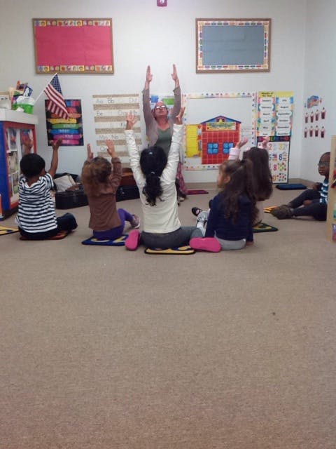 Person leading children in a classroom with arms raised