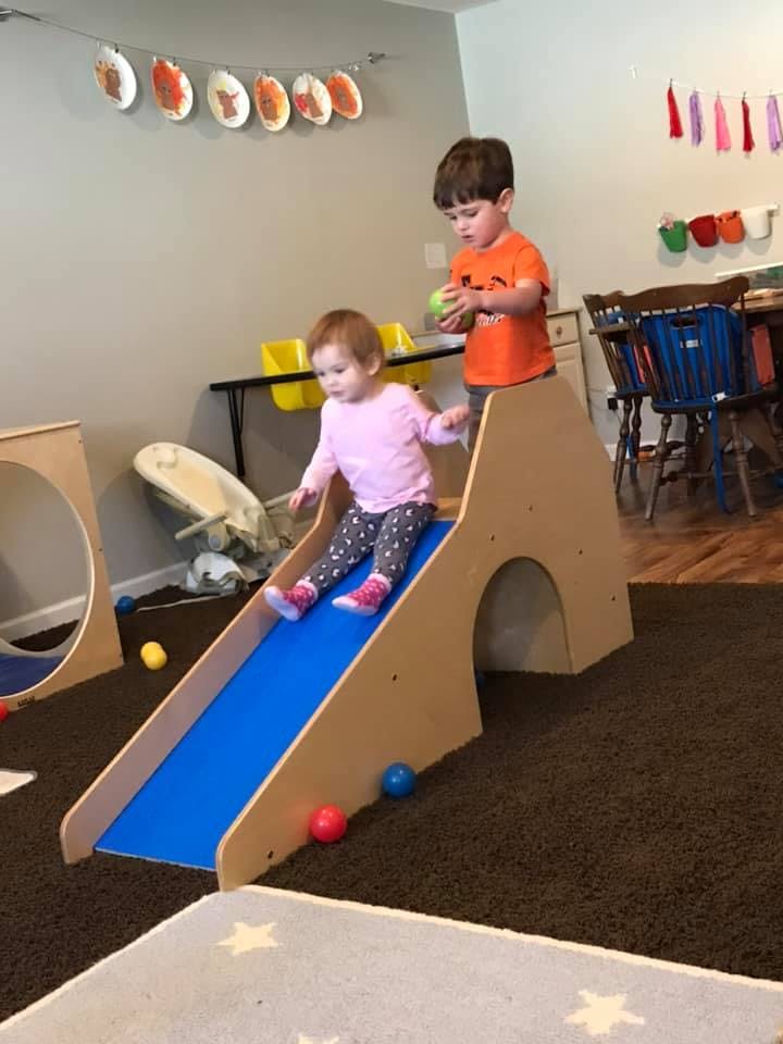 Child sliding down a blue slide while another child stands nearby holding a ball