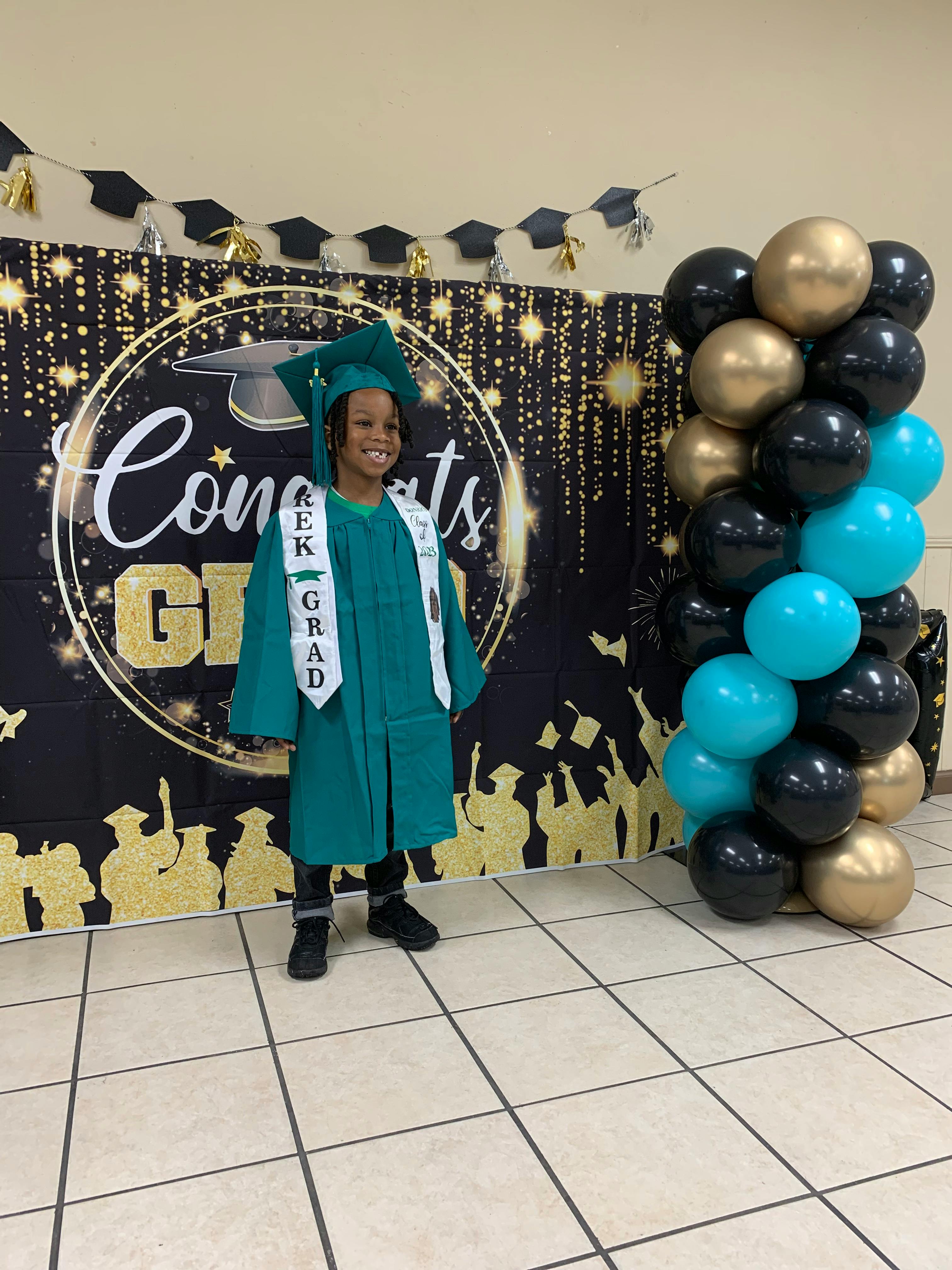 Child in a green graduation cap and gown standing in front of a banner
